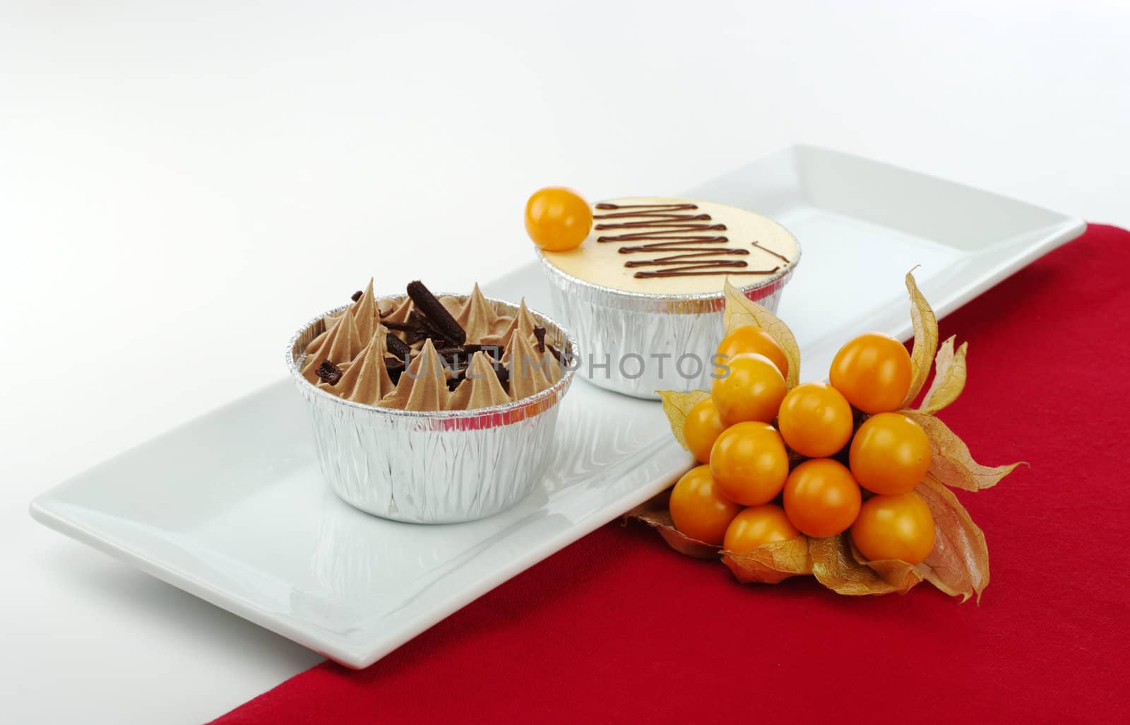 Two small cakes in tinfoil cups on rectangular white plate with physalis and red table mat (Selective Focus)