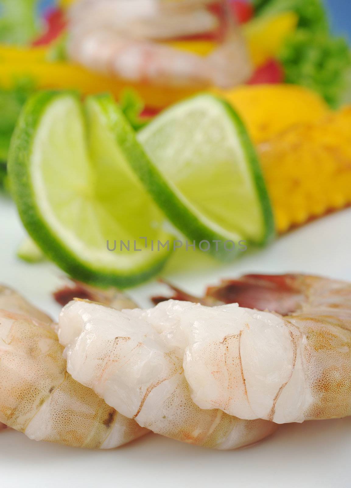 Closeup of raw king prawns with lime slices and lettuce on white plate with blue background  (Selective Focus)