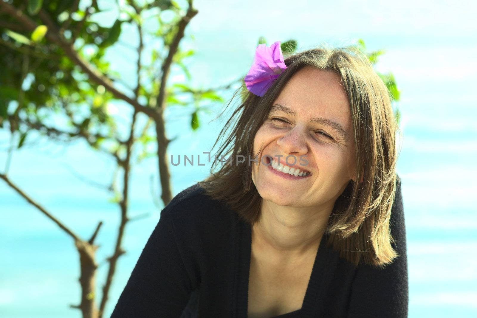 Smiling young woman with purple flower in her hair on a sunny day on the coast (Selective Focus, Focus on the left eye)
