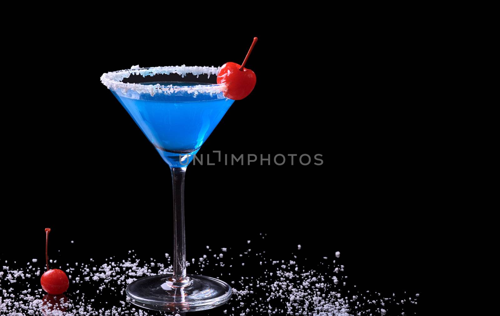 Blue Curacao drink in a glass with coconut flake rim and a maraschino cherry surrounded by coconut flakes and a maraschino cherry photographed on black (Selective Focus, Focus on the front of the glass and the cherry on the rim)