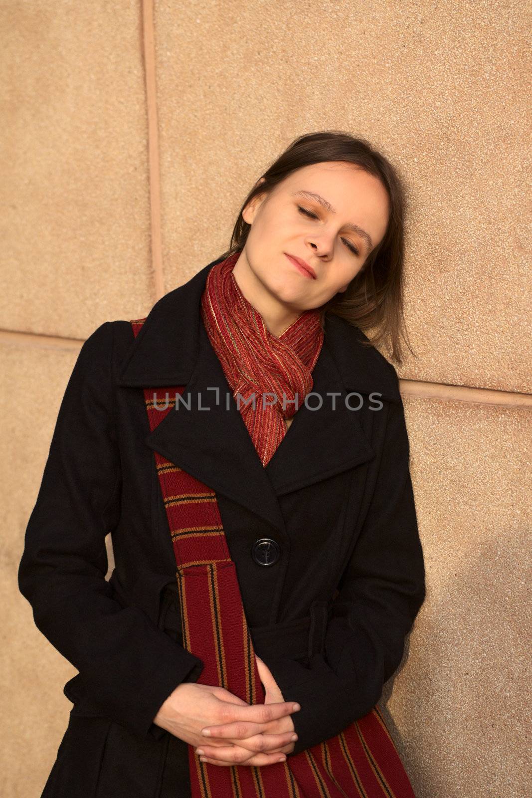 Young woman leaning against a column with closed eyes enjoying the warmth of the setting sun (Selective Focus, Focus on the face)