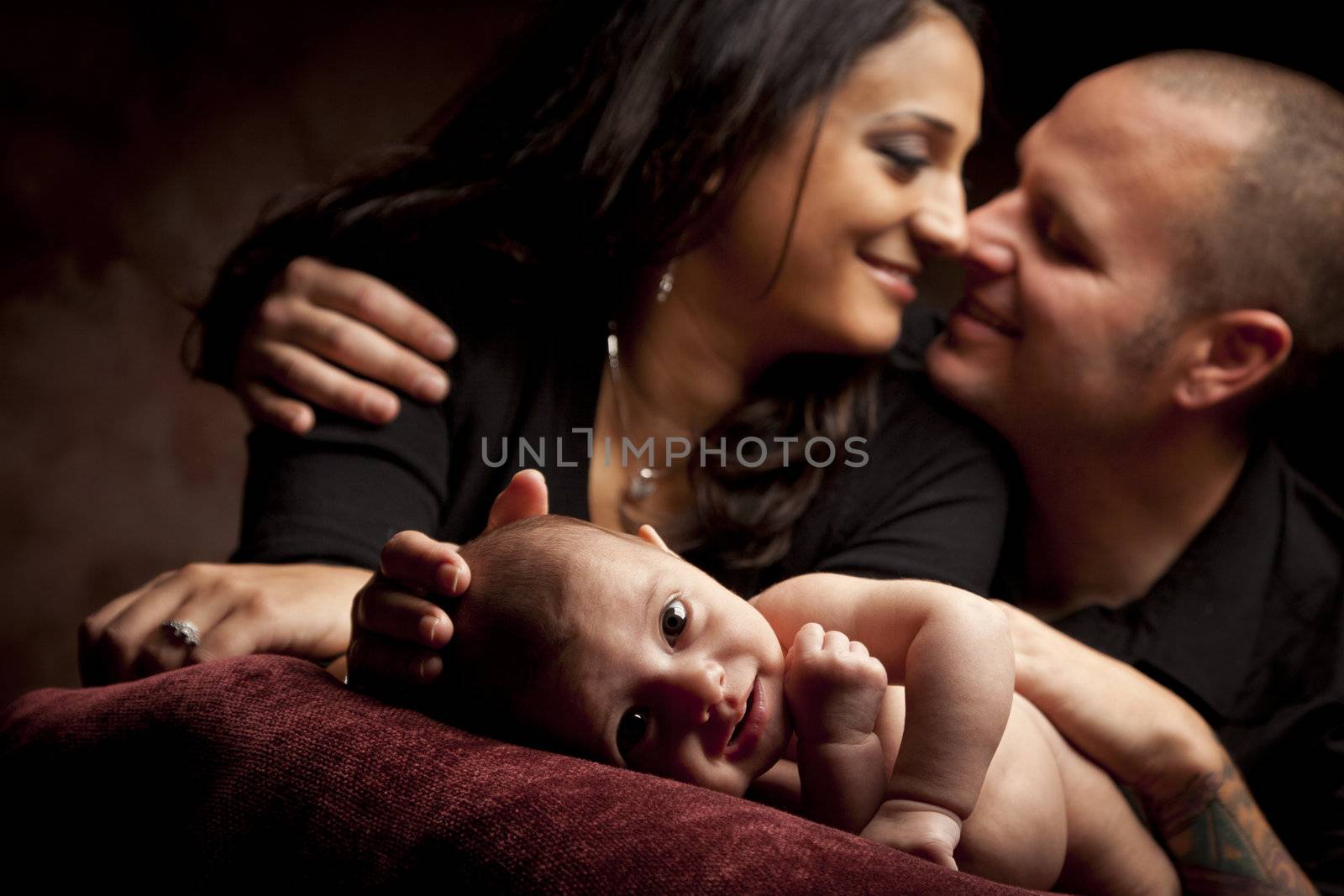 Mixed Race Couple Lovingly Look On While Baby Lays on Pillow on a Dark Background.