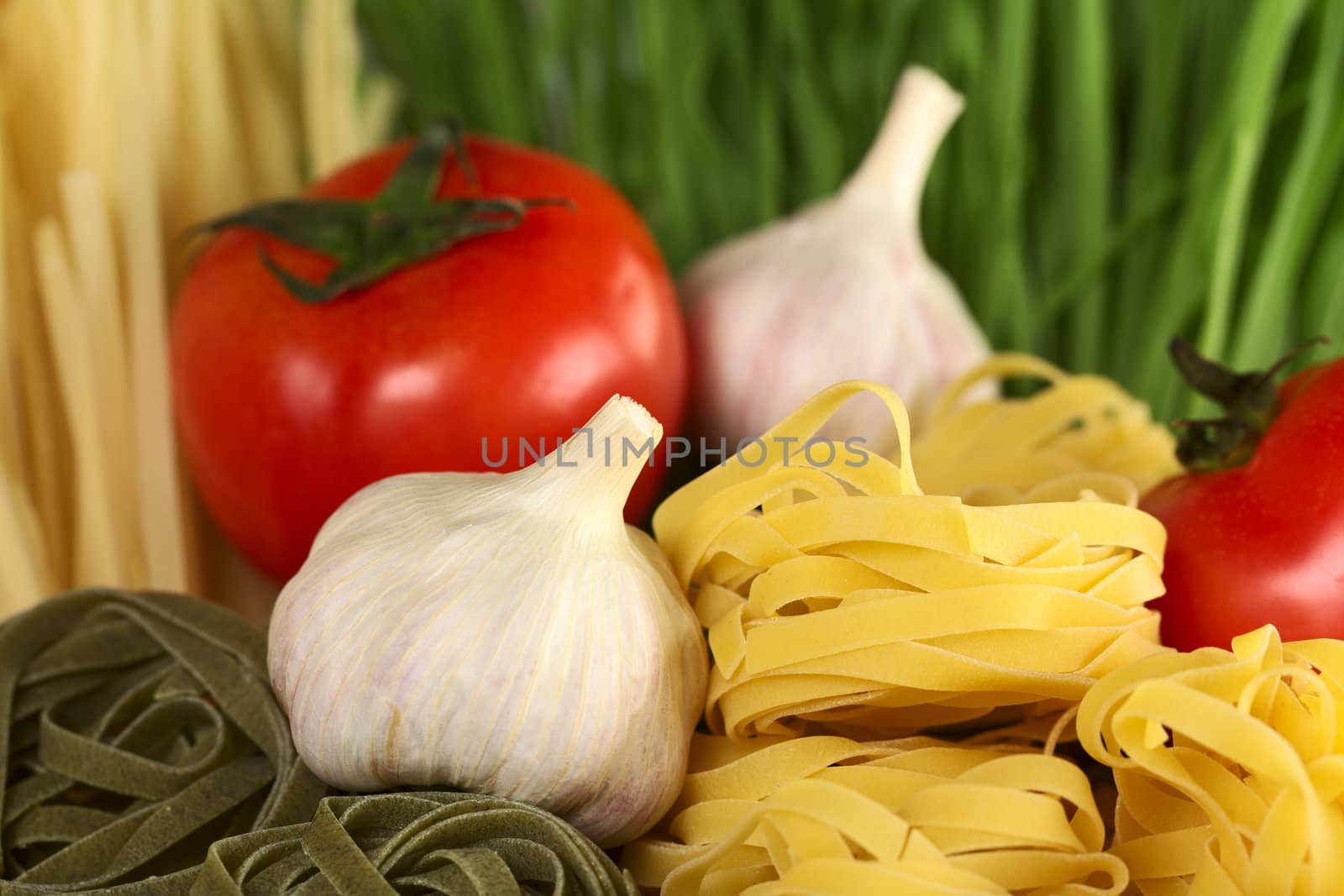 Raw tagliatelle paglia e fieno (straw and hay) with raw garlic bulb and globe tomato (Selective Focus, Focus on the front of the upper yellow tagliatelle pile and parts of the garlic bulb beside)