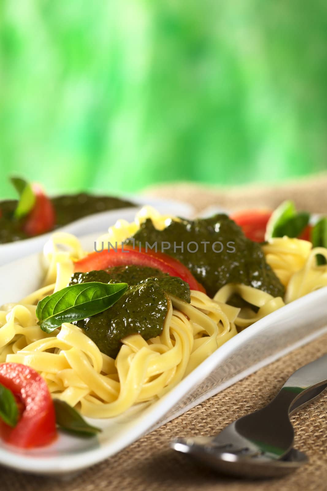 Yellow tagliatelle with fresh pesto made of basil and garlic and garnished with tomato slices and basil leaves (Selective Focus, Focus on the basil leaf on the front of the pesto)
