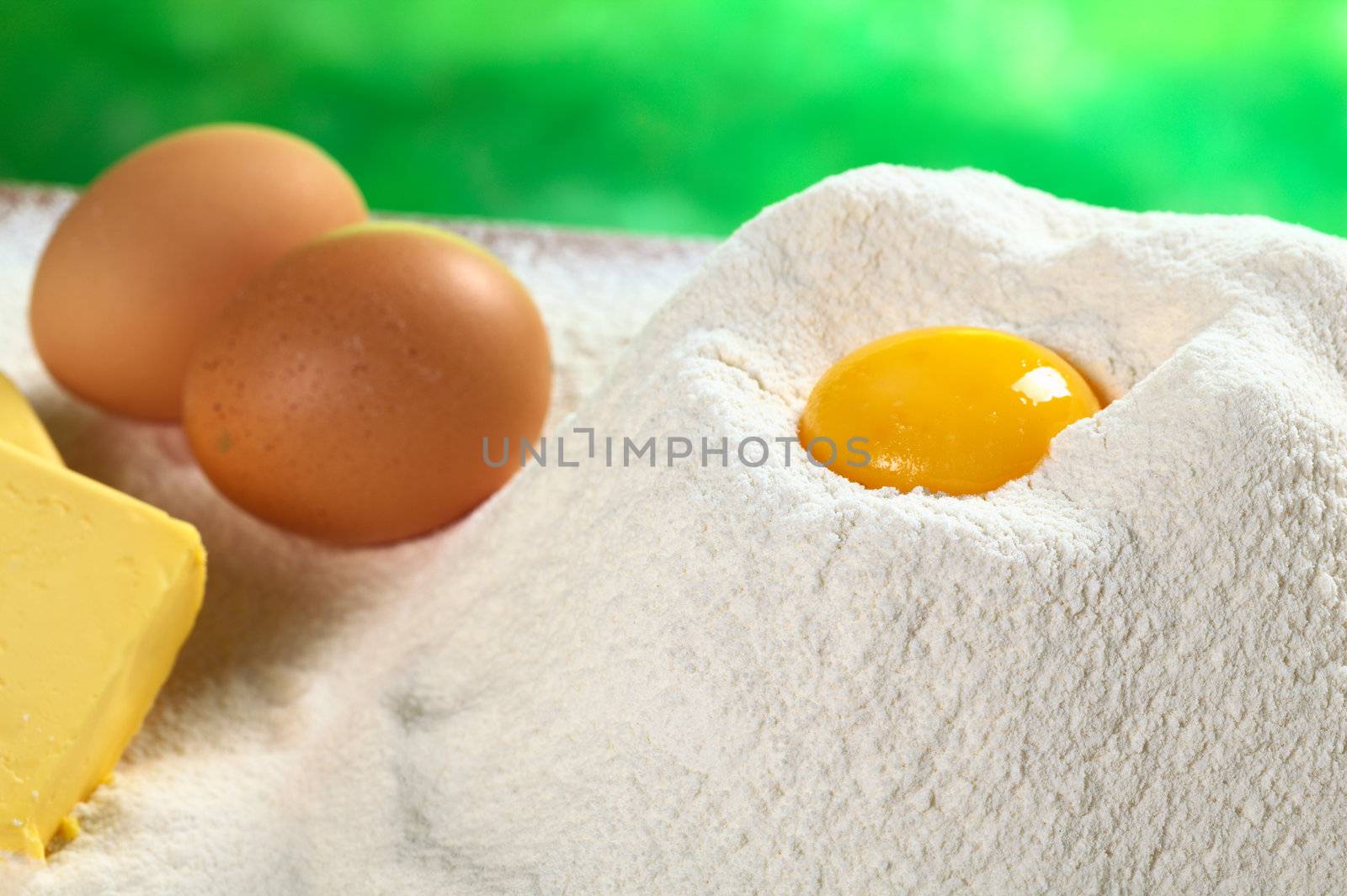 Flour with egg yolk in the middle with eggs and butter on the side (Selective Focus, Focus on the front of the flour pile and the front of the egg yolk)