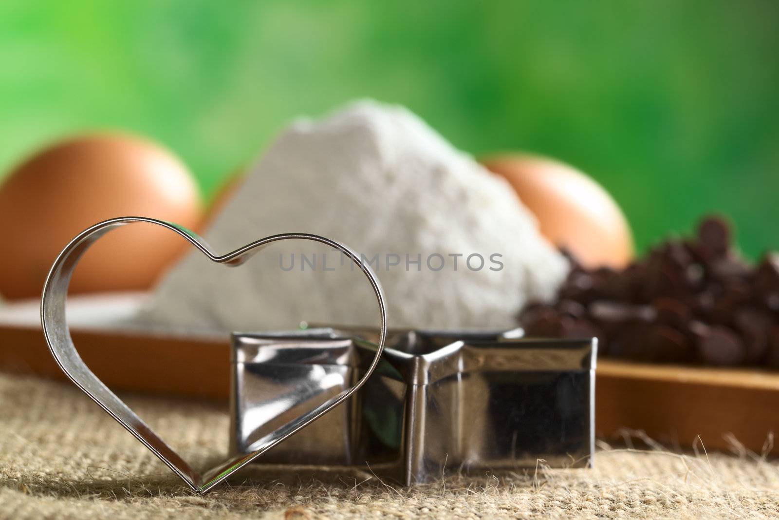 Heart-shaped cookie cutter leaning against a star-shaped cutter with baking ingredients such as wheat flour, chocolate chips and eggs in the back (Selective Focus, Focus on the heart-shaped cookie cutter)