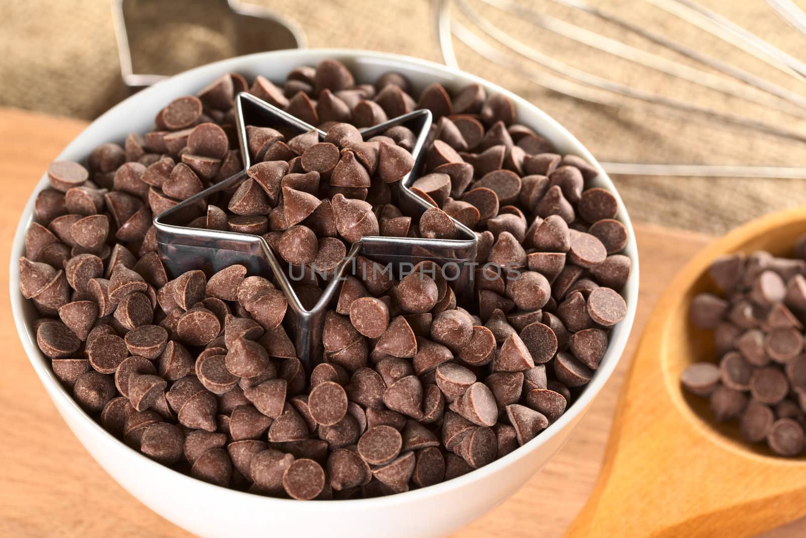 Star-shaped cookie cutter with chocolate chips in a bowl surrounded by baking utensils (Selective Focus, Focus on the front of the cutter and the chocolate chips around)