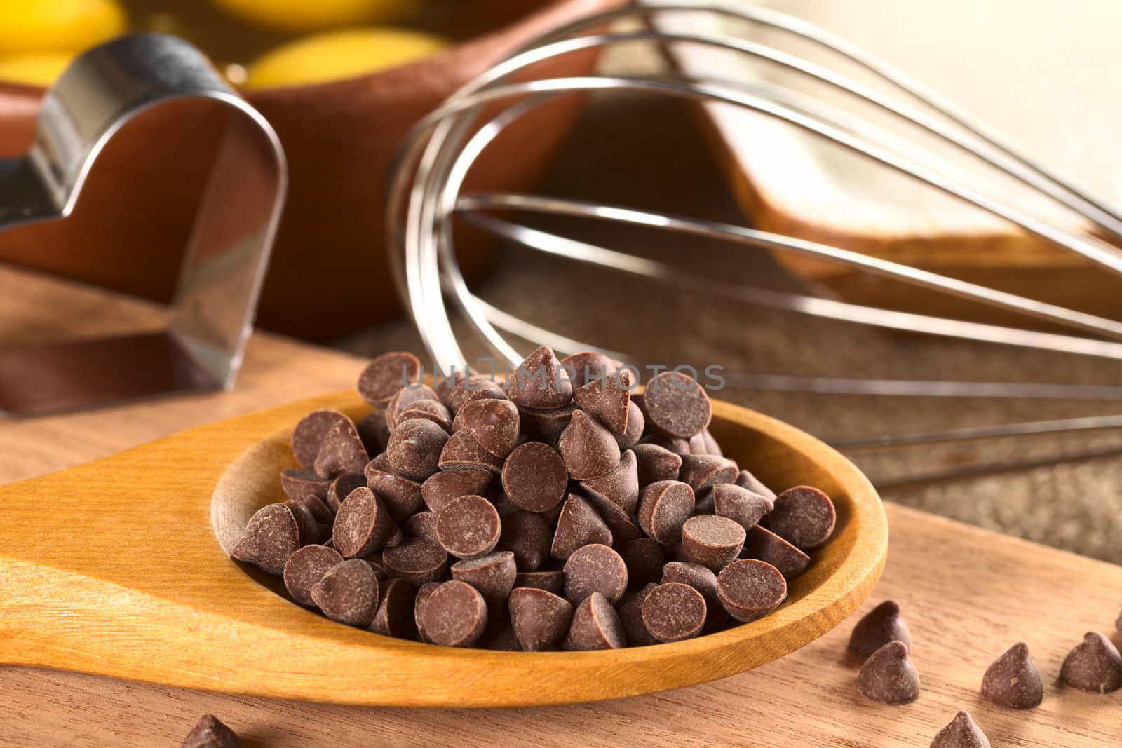 Chocolate chips on wooden spoon with beater, cutter, egg and flour in the back (Selective Focus, Focus on the chocolate chips on top and the ones in the same plane)