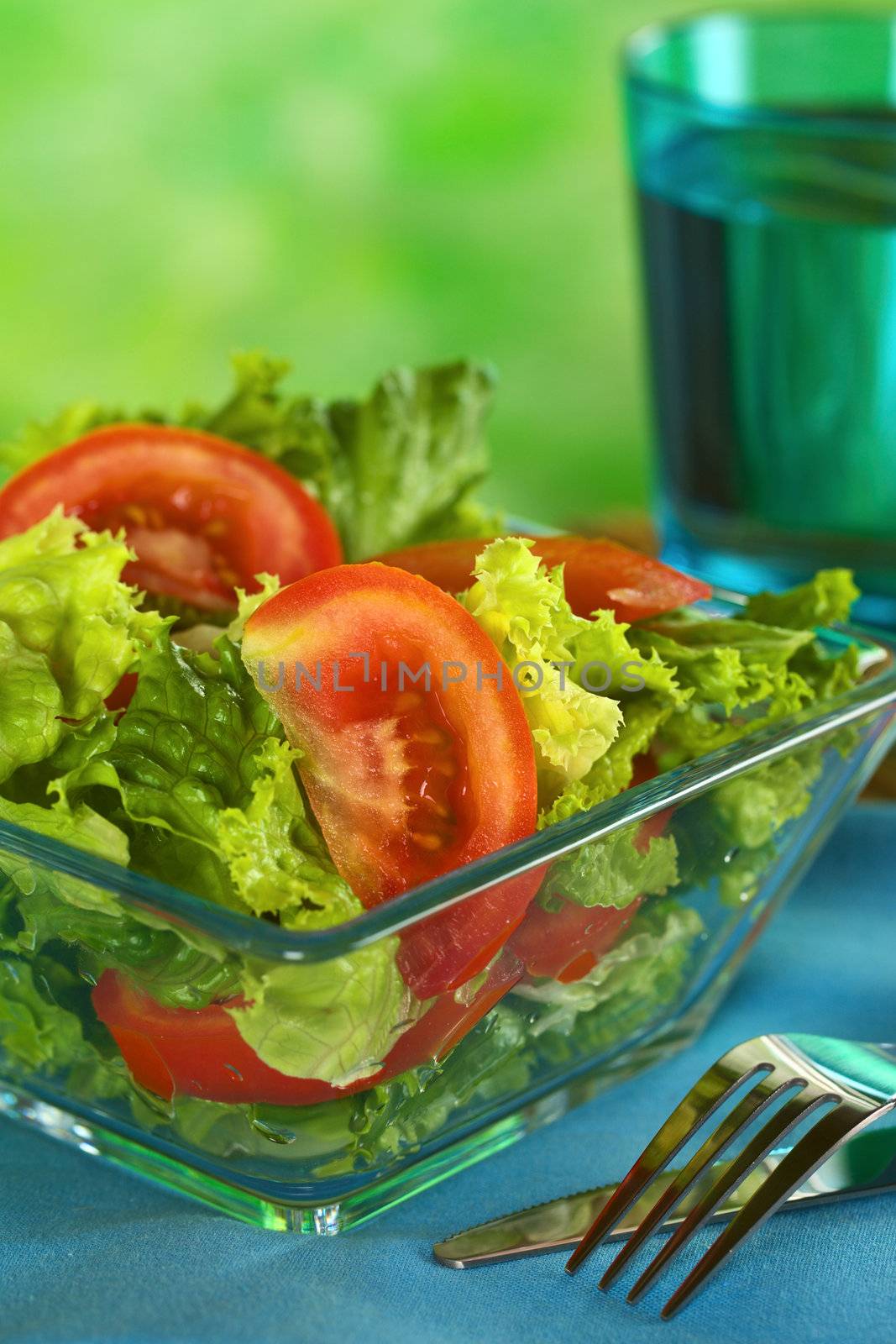Fresh salad of lettuce and tomato with a glass of water in the back (Selective Focus, Focus on the tomato slice in the front) 