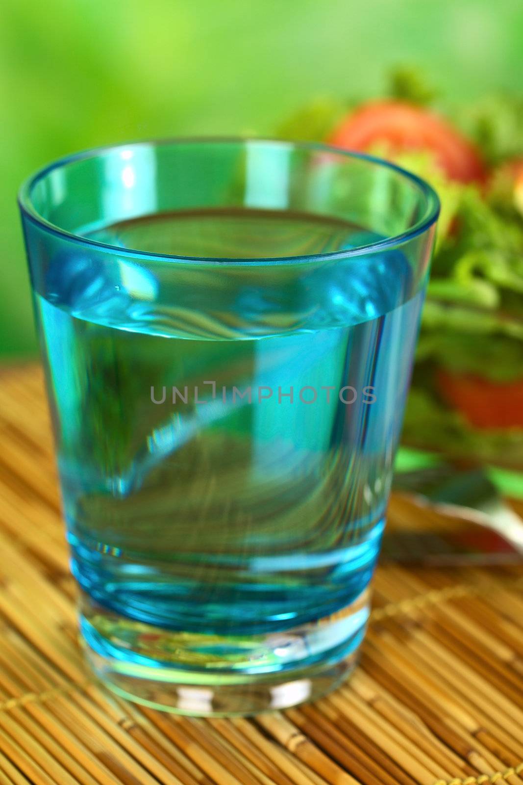 Glass of still mineral water with fresh salad in the back (Selective Focus, Focus on the front rim of the glass)