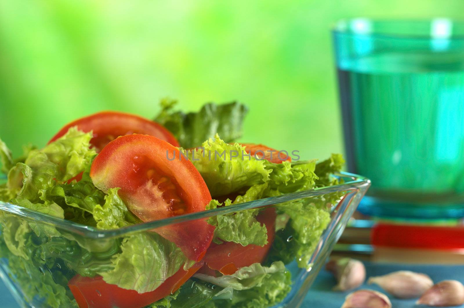 Fresh salad of lettuce and tomato with garlic and a glass of water in the back (Selective Focus, Focus on the tomato slice in the front) 