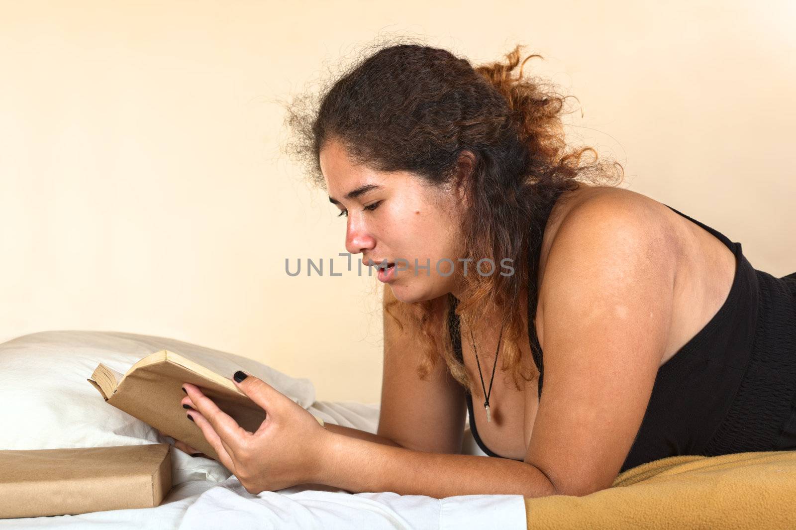 Young Peruvian woman lying on bed and reading a book (Selective Focus, Focus on the left eye of the woman)