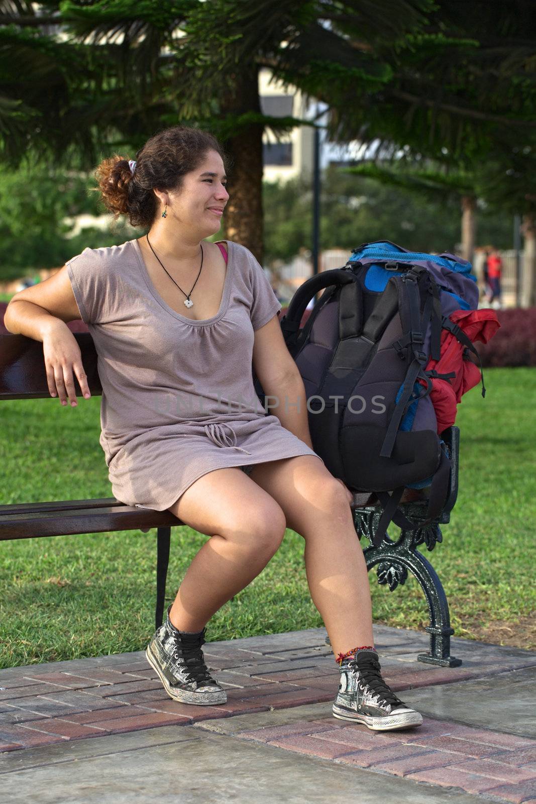 Young smiling Peruvian woman with backpack sitting on a bench in park (Selective Focus, Focus on the face of the woman)