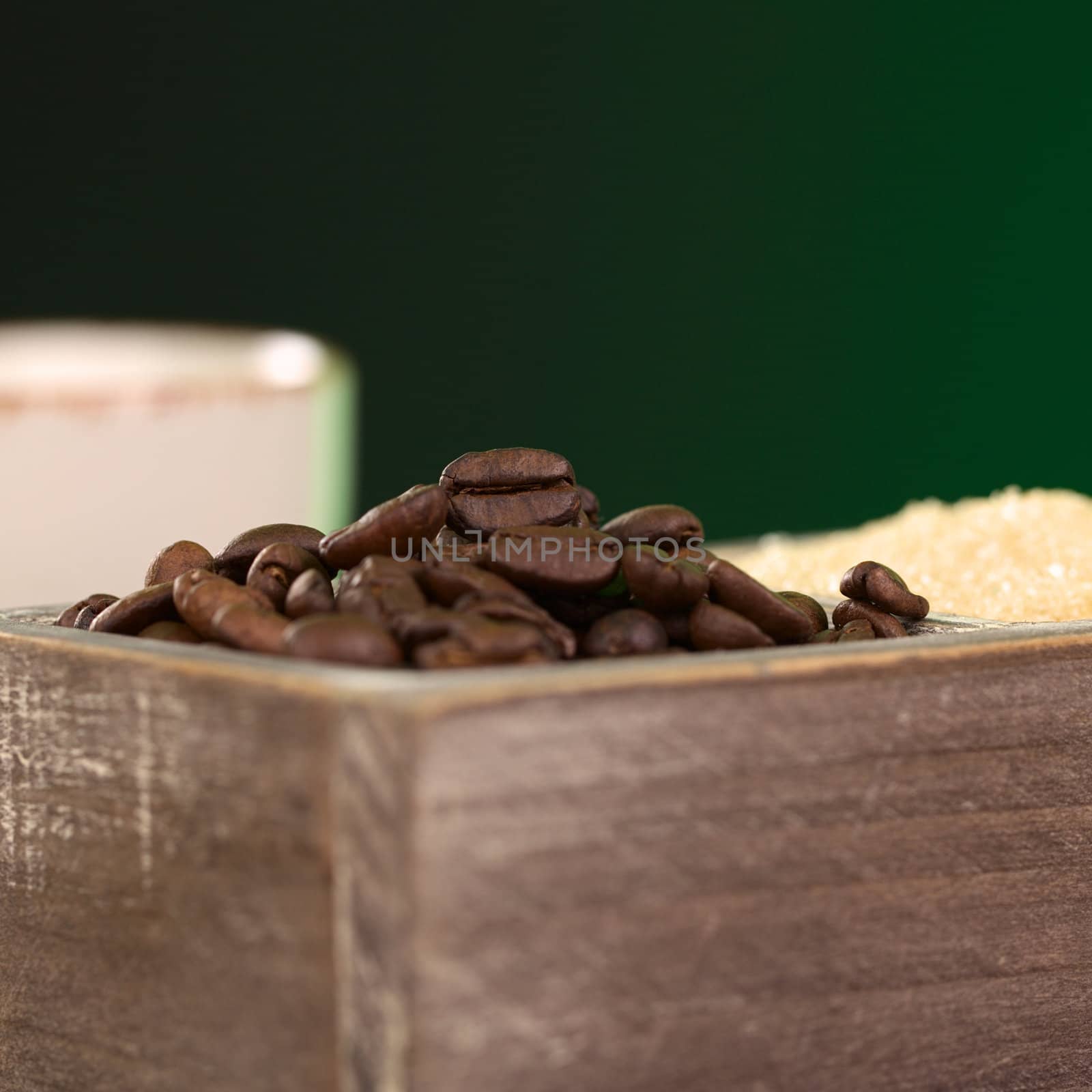 Roasted coffee beans and brown sugar in wooden container with coffee cup in the back and dark green background (Selective Focus, Focus on the coffee bean on the top of the pile)