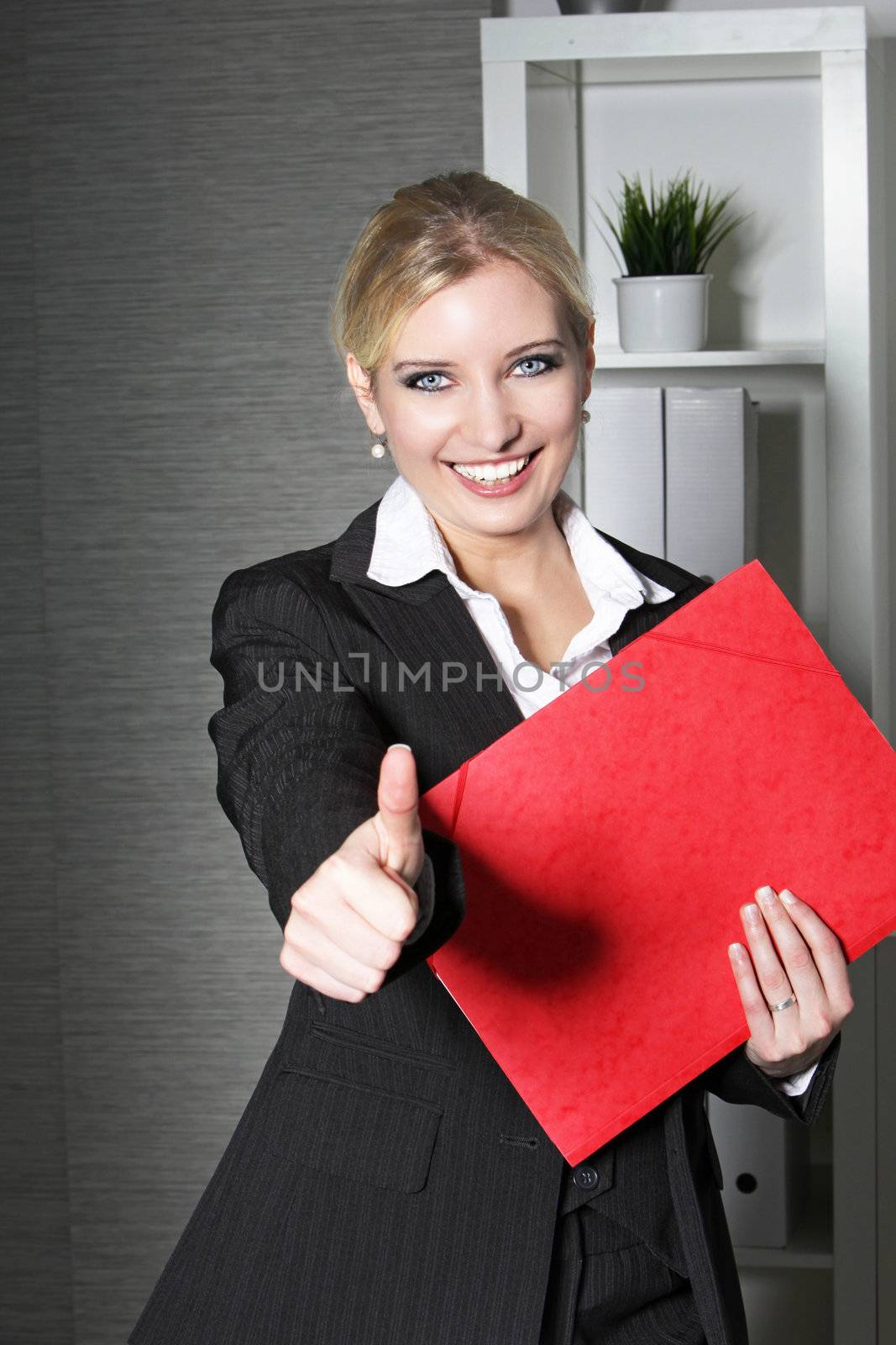 Beautiful stylish young female office worker holding a red folder giving a thumbs up gesture with a big cheerful smile