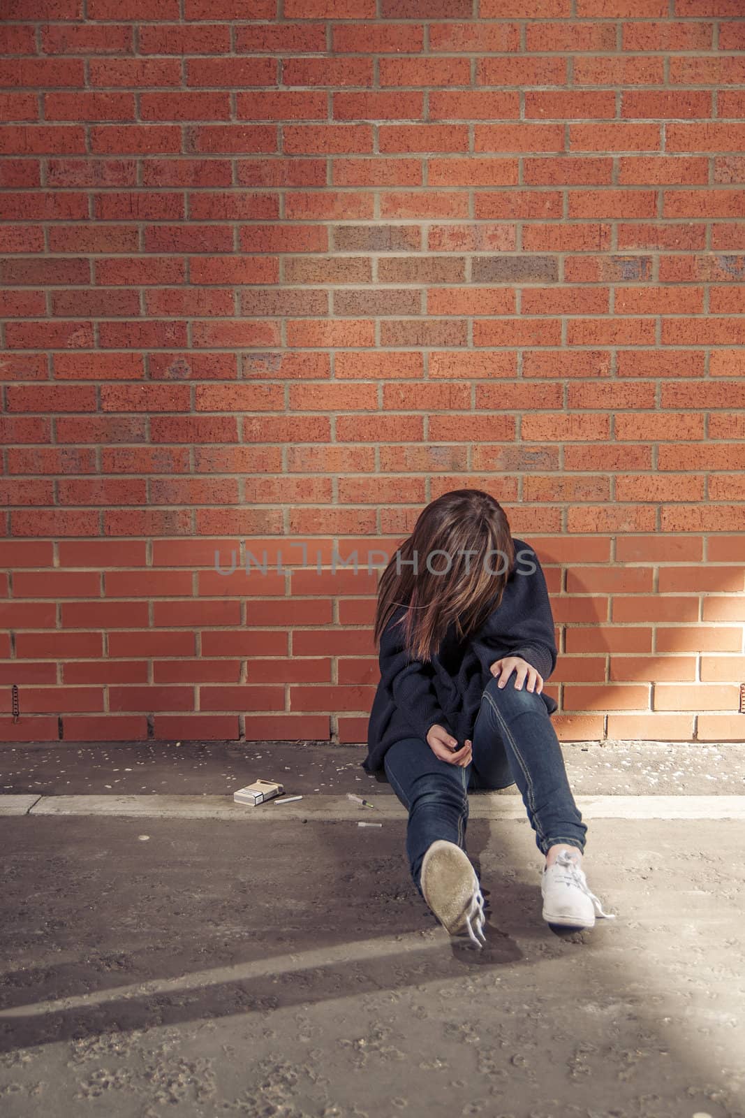 Addicted, sad young woman sitting against the brick wall with syringe and cigarettes beside.  Vertical.