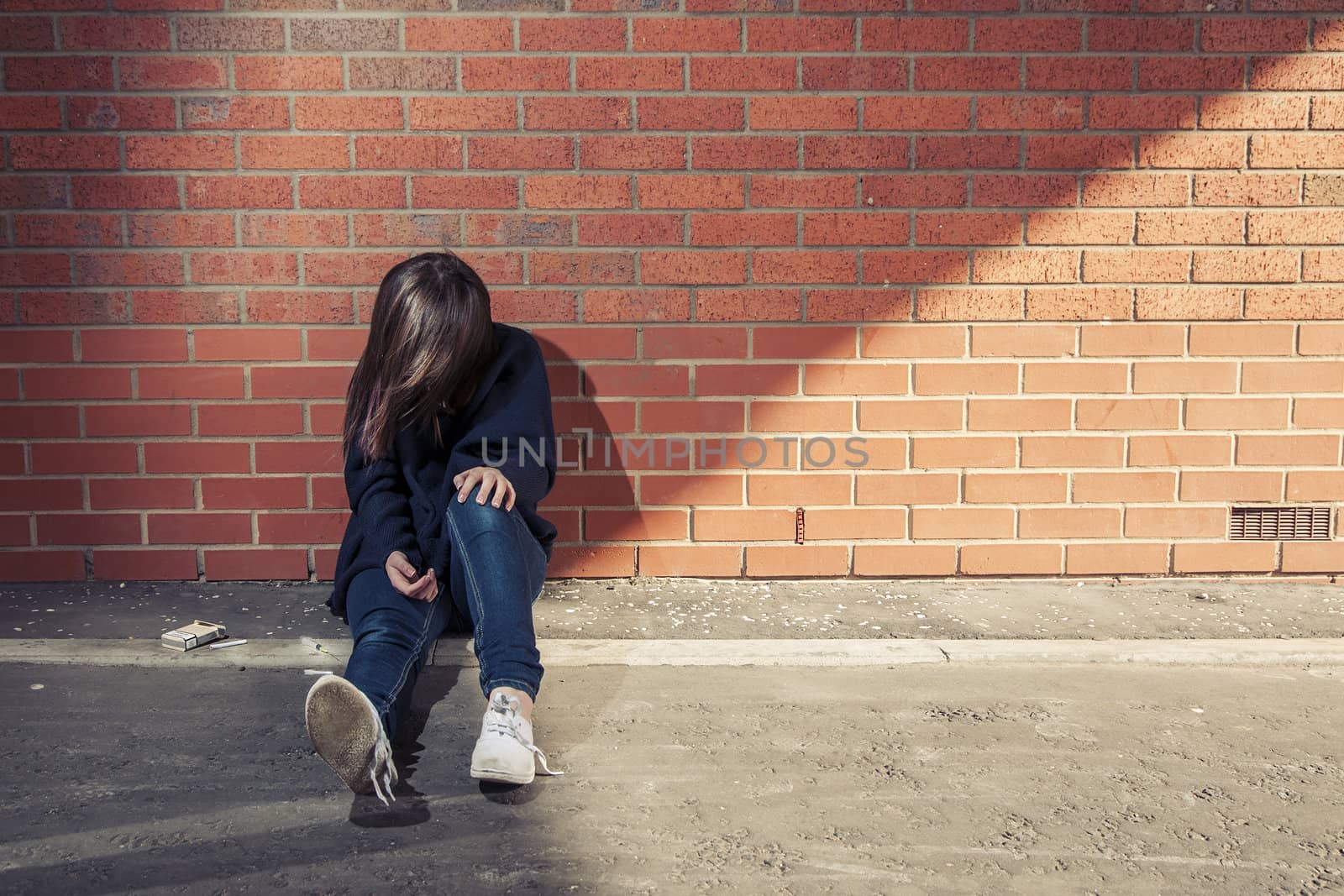 Addicted, sad young woman sitting against the brick wall with syringe and cigarettes beside.  Horizontal.