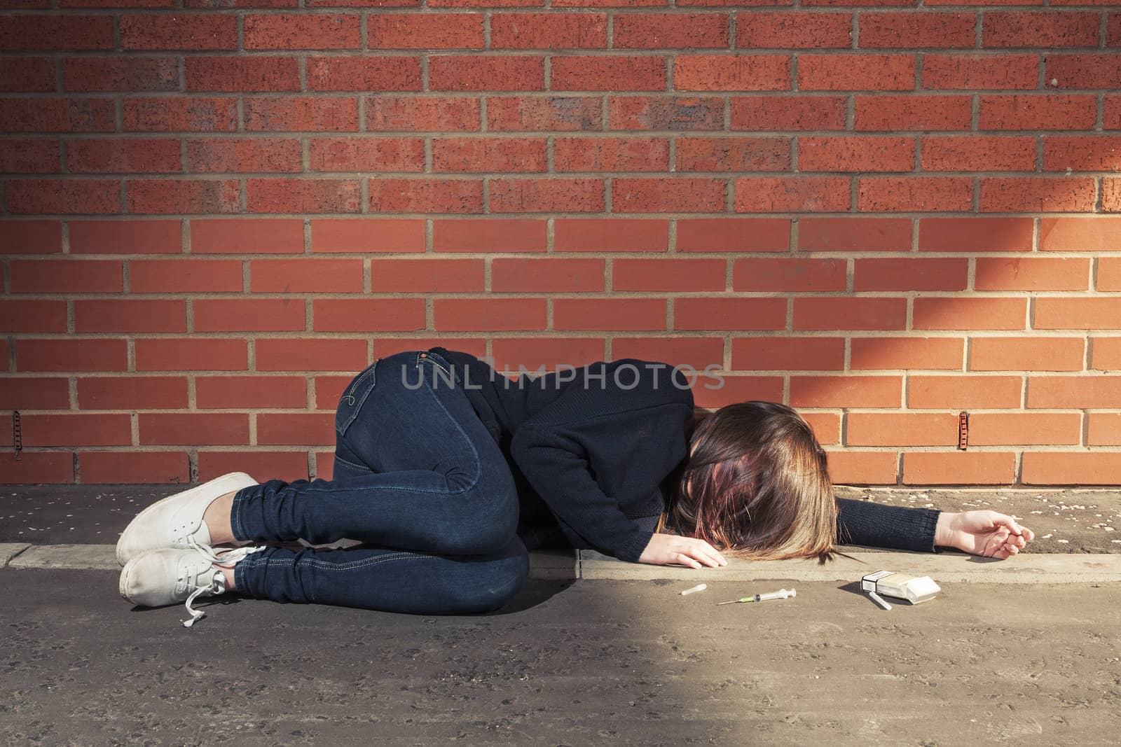 Addicted, sad young woman lying against the brick wall with syringe and cigarettes beside.  Horizontal.
