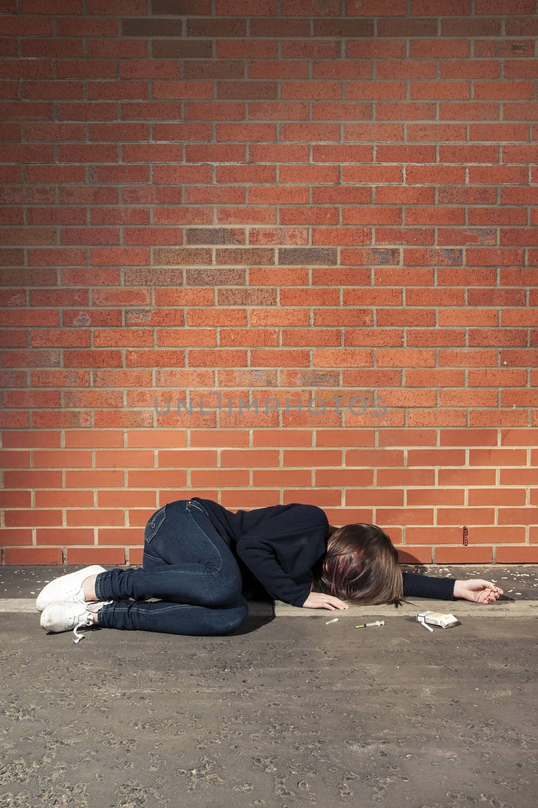 Addicted, sad young woman lying against the brick wall with syringe and cigarettes beside. Vertical.