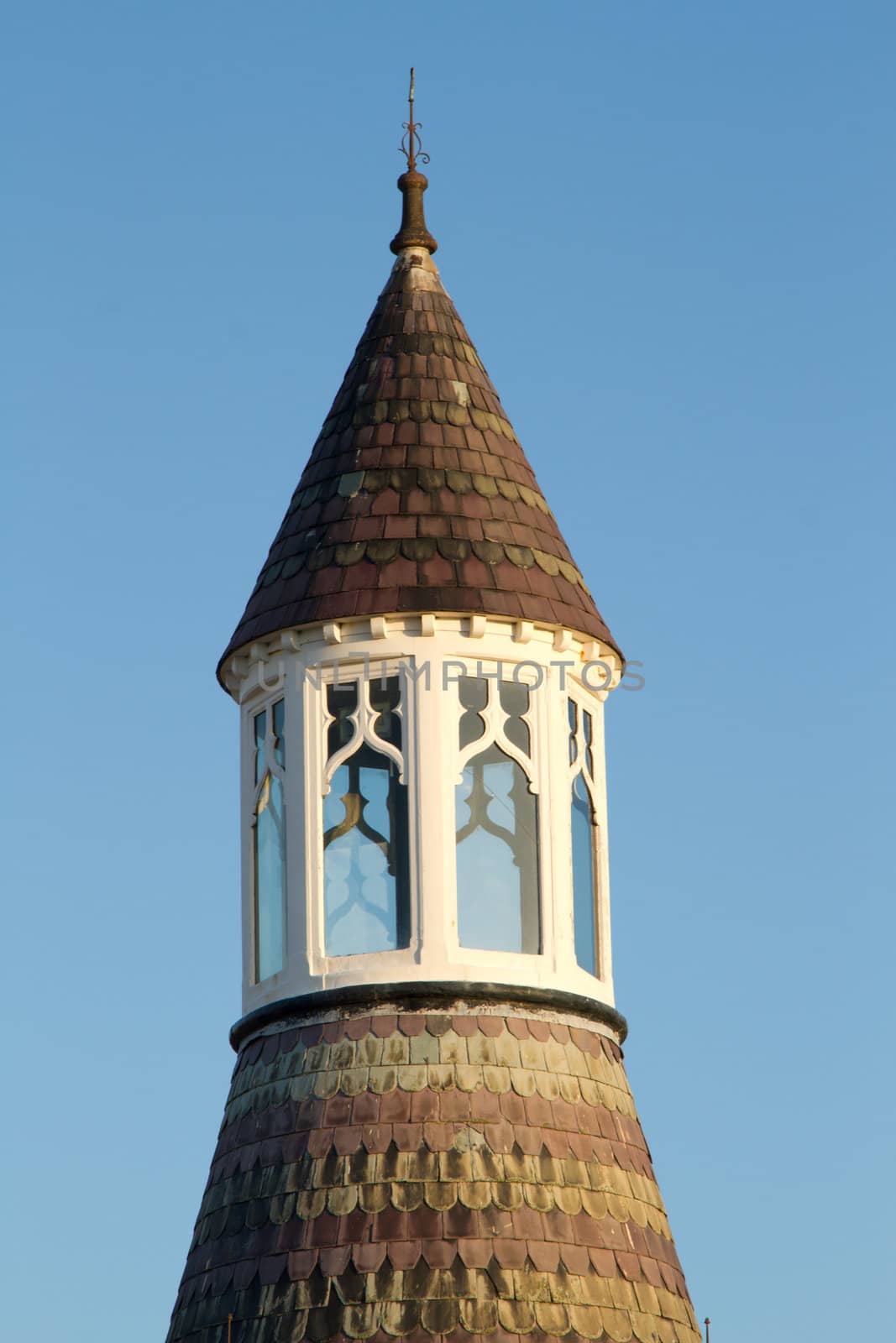 A tower, turret, with patterned red and green slate tiles a windowed viewing area and lightening conductor on the top against a blue sky.