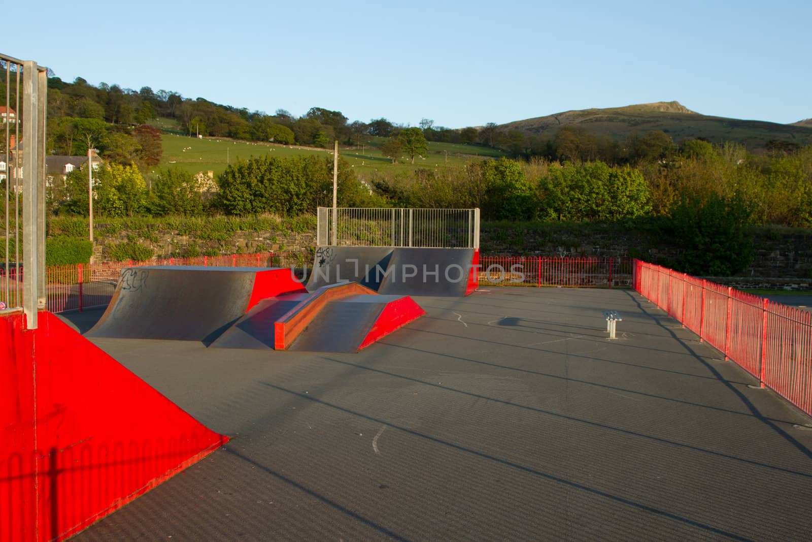 An outdoor skatepark with variuos ramps made from metal painted red with green fields in the background.