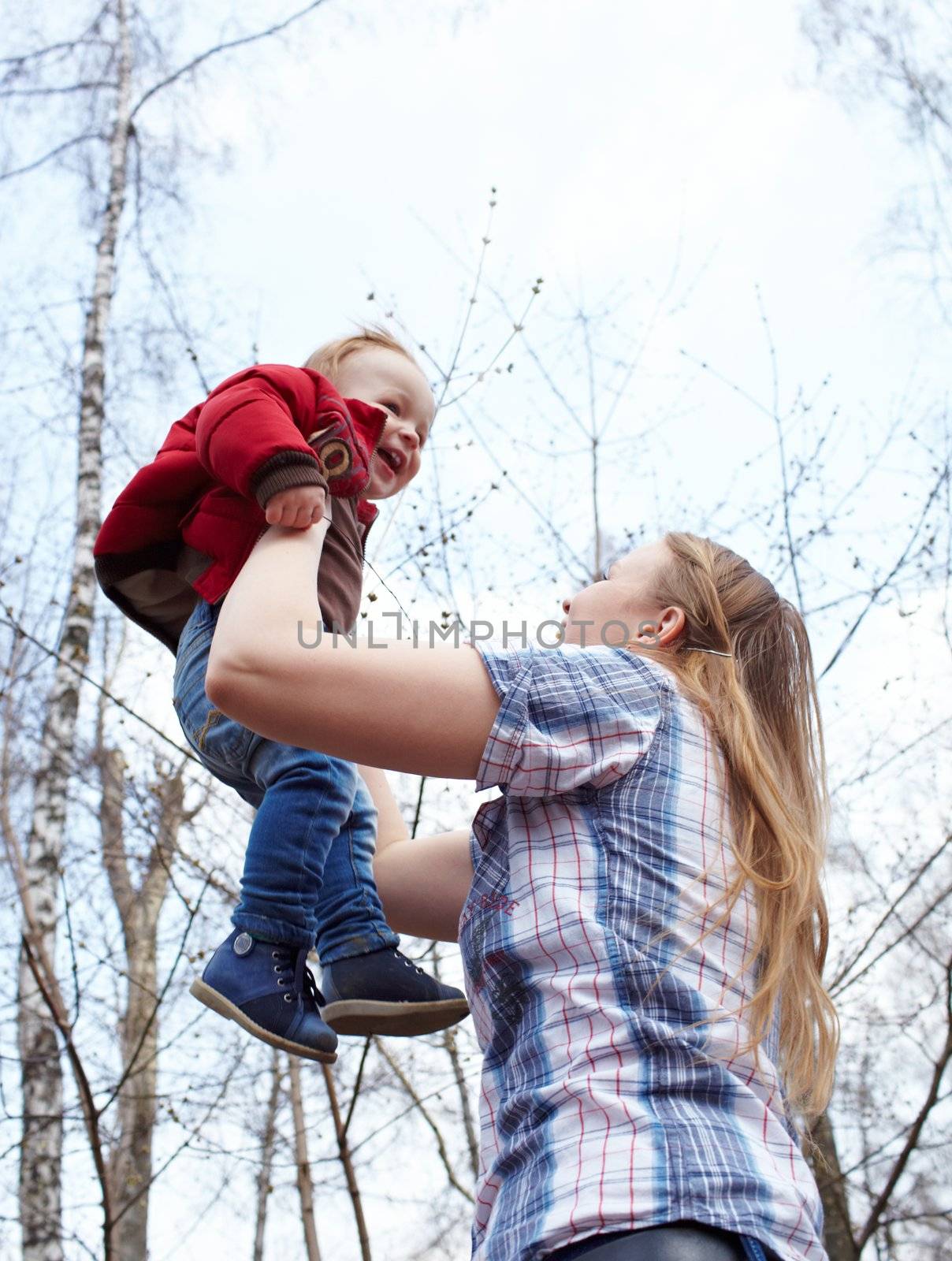 Happy young mother and her son laughing and playing together in the park on the sky background. This shot was made in the first day of spring.