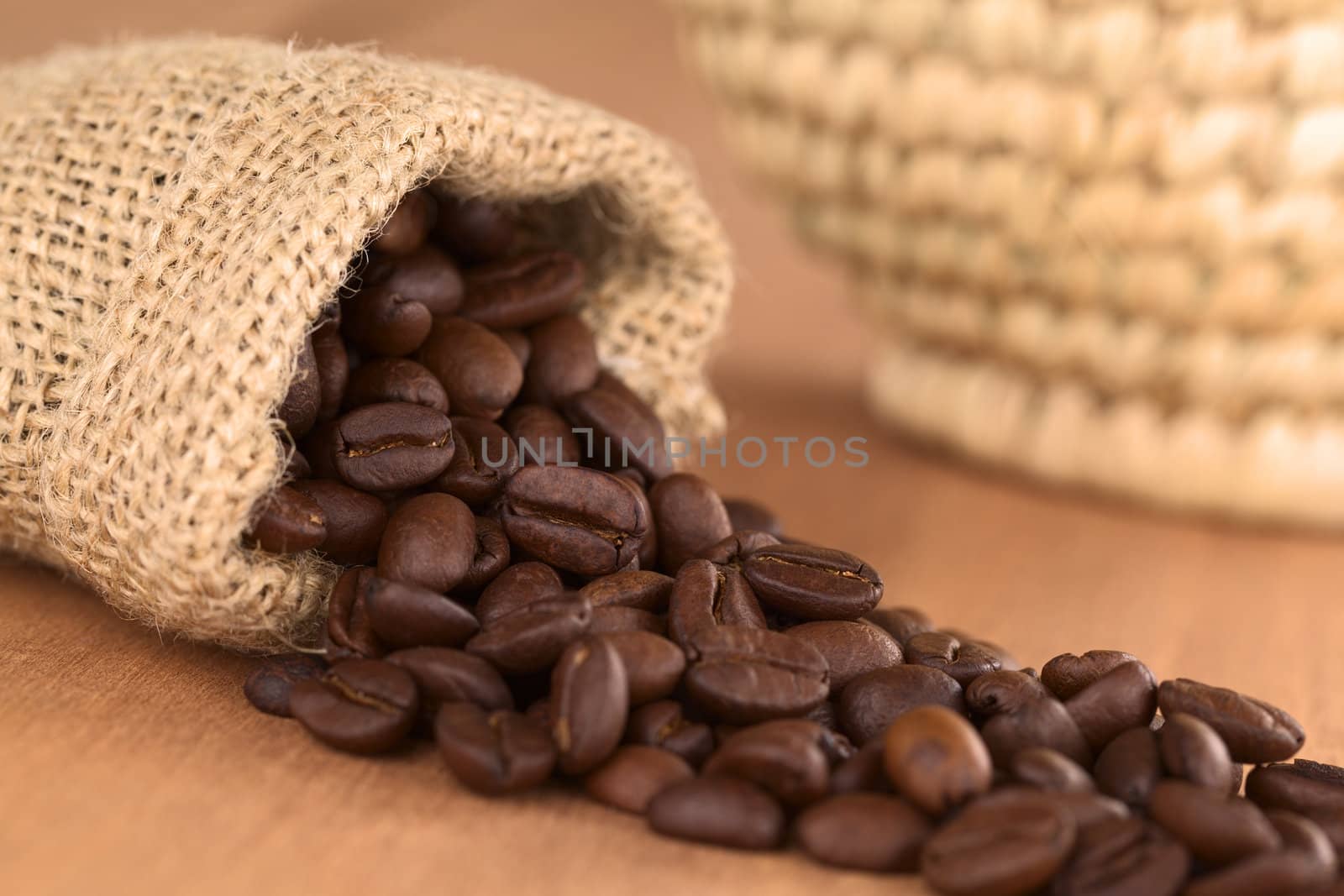 Roasted coffee beans in jute bag on wood with woven basket in the back (Selective Focus, Focus on the coffee beans turned to the front in the middle of the image) 