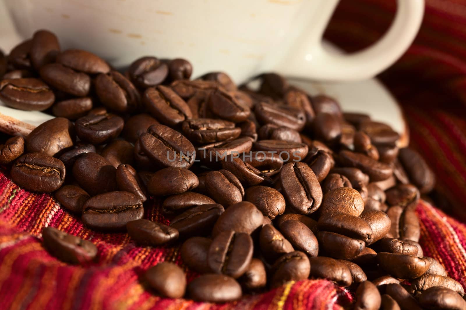 Roasted coffee beans on red cloth with coffee cup in the back (Selective Focus, Focus on the coffee beans in the middle of the image)