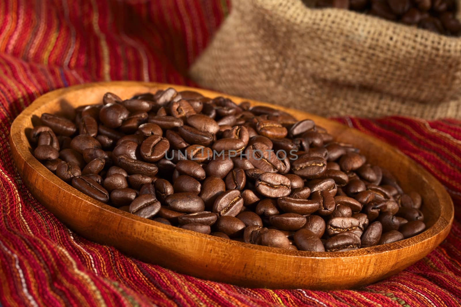 Roasted coffee beans on wooden plate on red cloth with jute bag in the back (Selective Focus, Focus one third into the coffee beans)