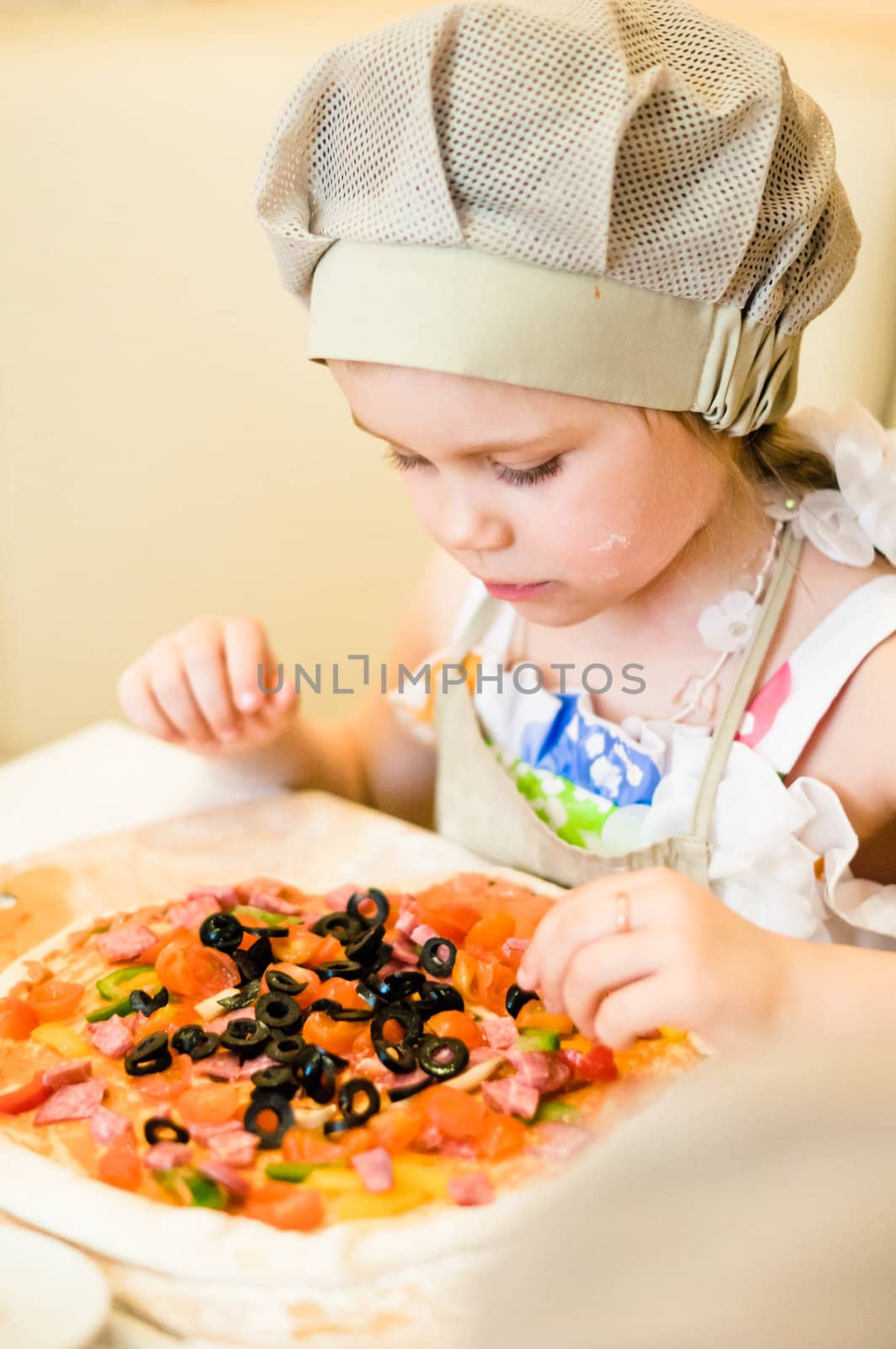 Little girl adding ingredients, vegetables and meat, in pizza