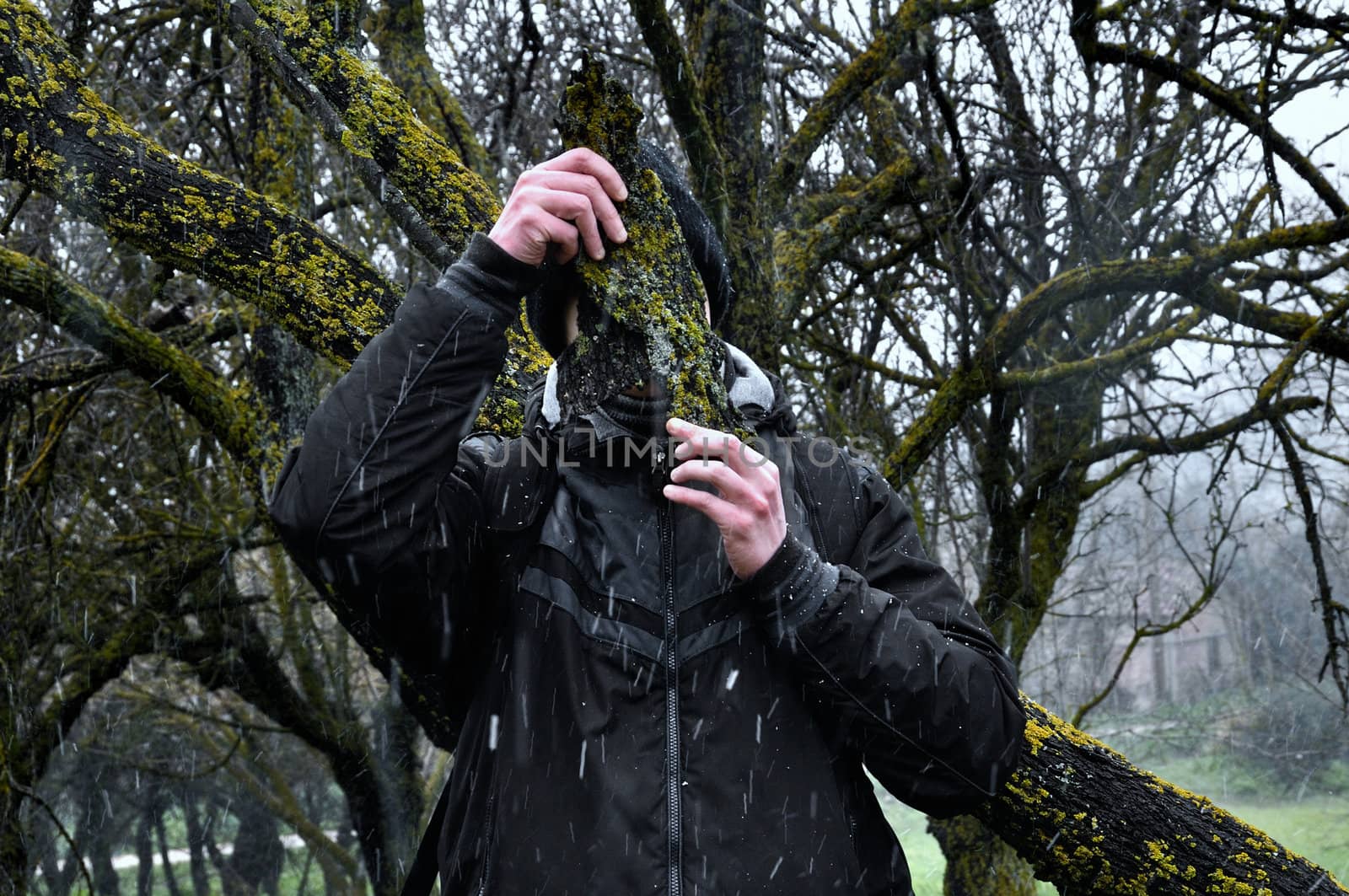 Man with bark peel mask under tree branches and falling snow in the woods.