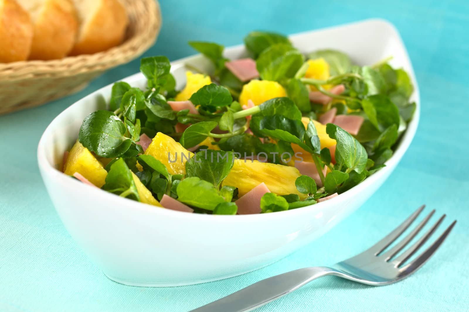 Fresh watercress, pineapple and ham salad in elongate white bowl with baguette slices in the back (Selective Focus, Focus on the two pineapple pieces one third into the salad)