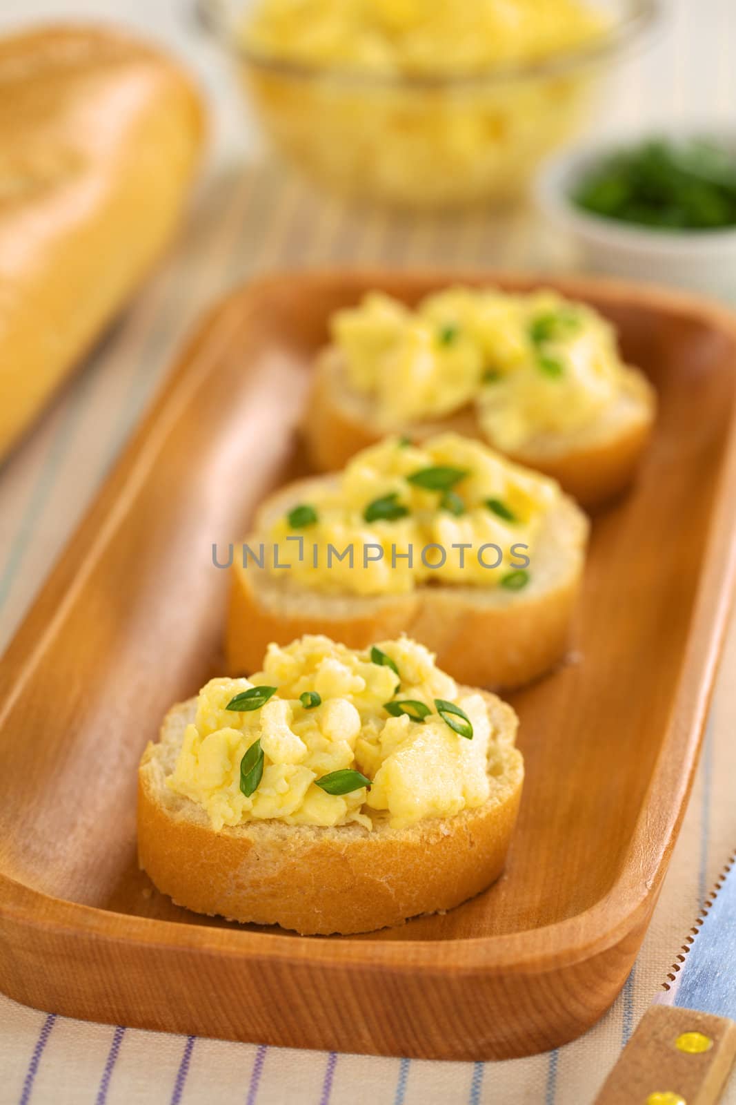 Scrambled eggs on baguette slices with green onion on elongate wooden plate (Selective Focus, Focus on the front of the first sandwich) 