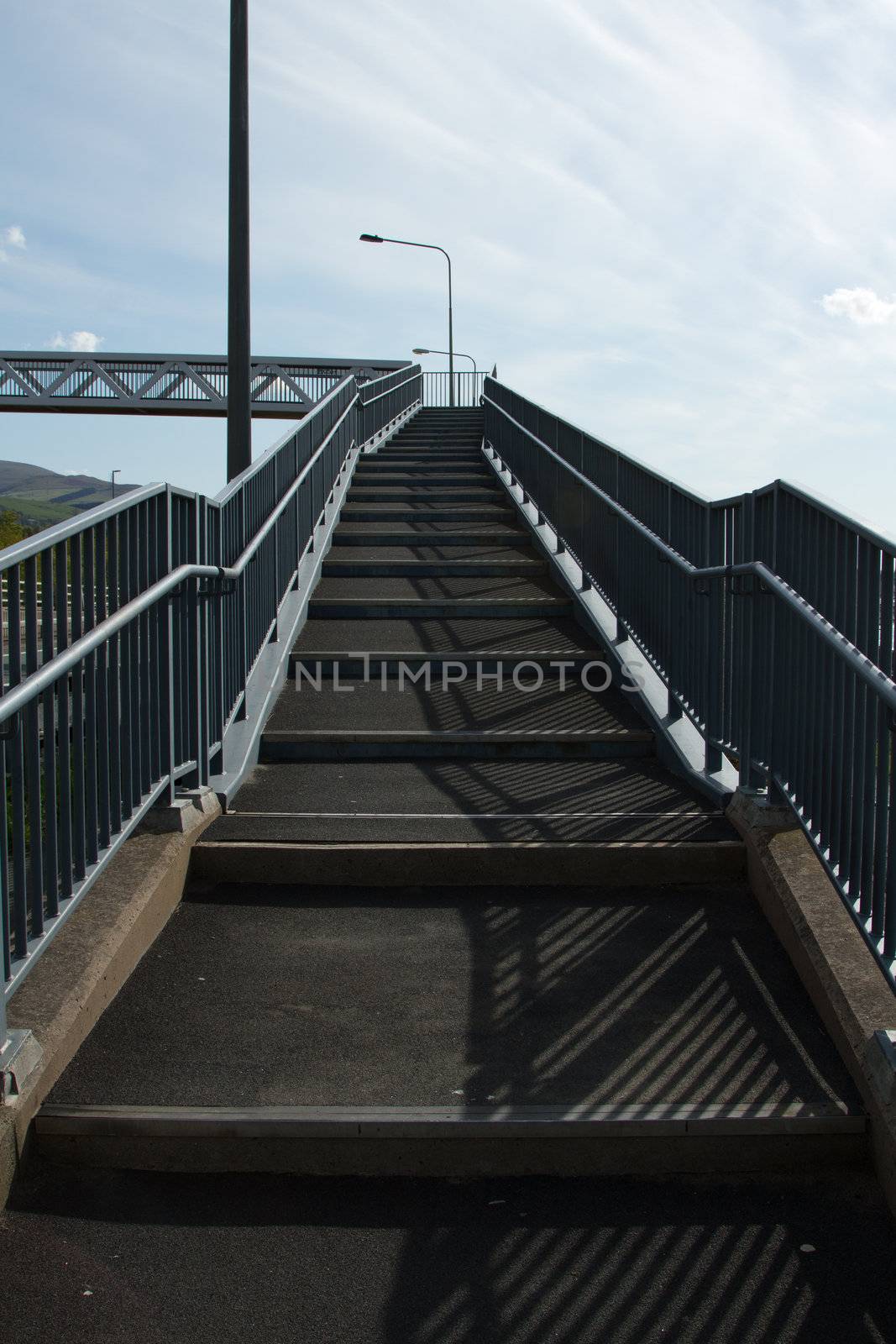 Steps leading to a foot bridge with metal railings and  lampposts with a blue sky and whispy clouds in the background.