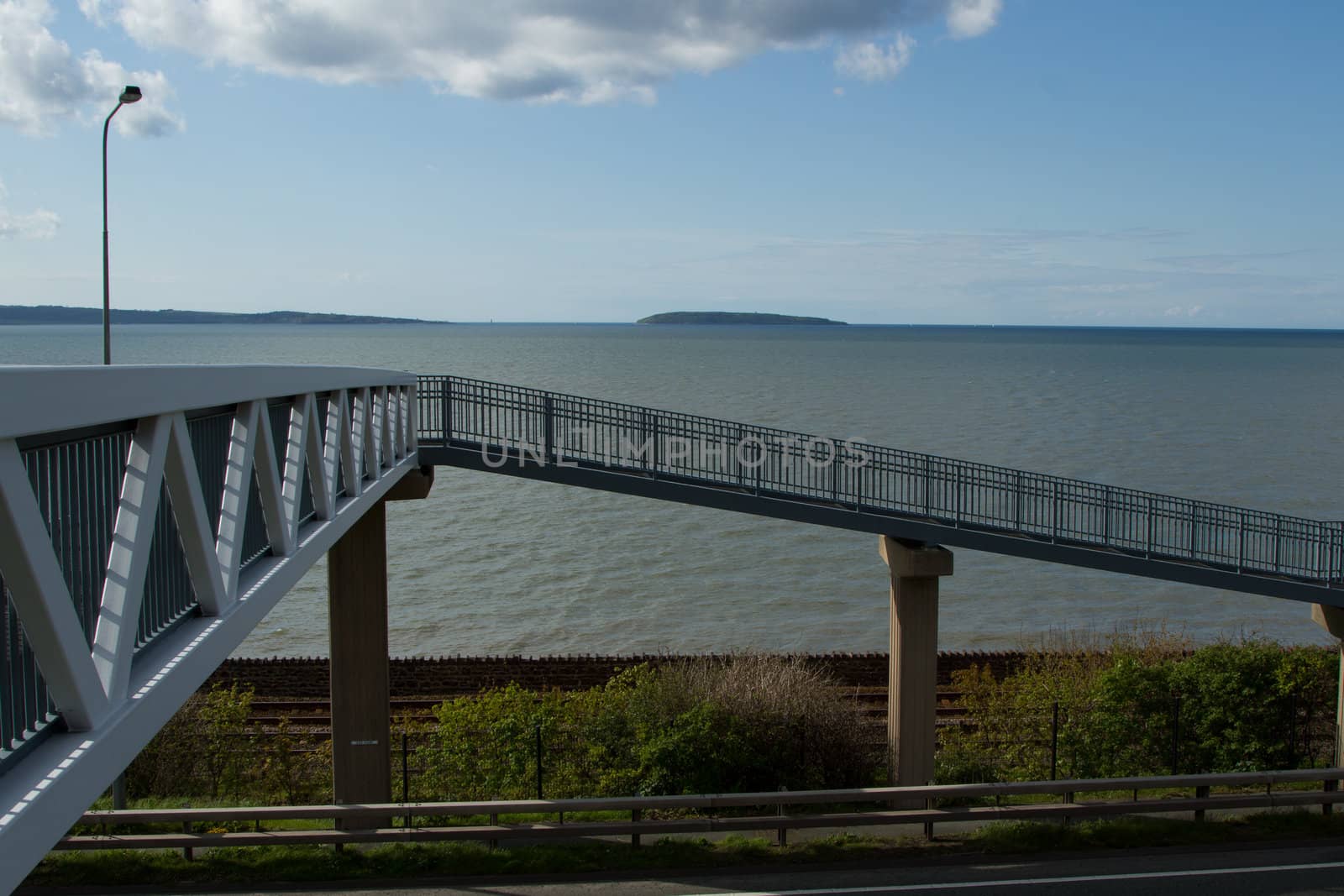 A metal footbridge leads to a ramp supported by concrete pillars with the sea, island, blue sky and cloud in the background.