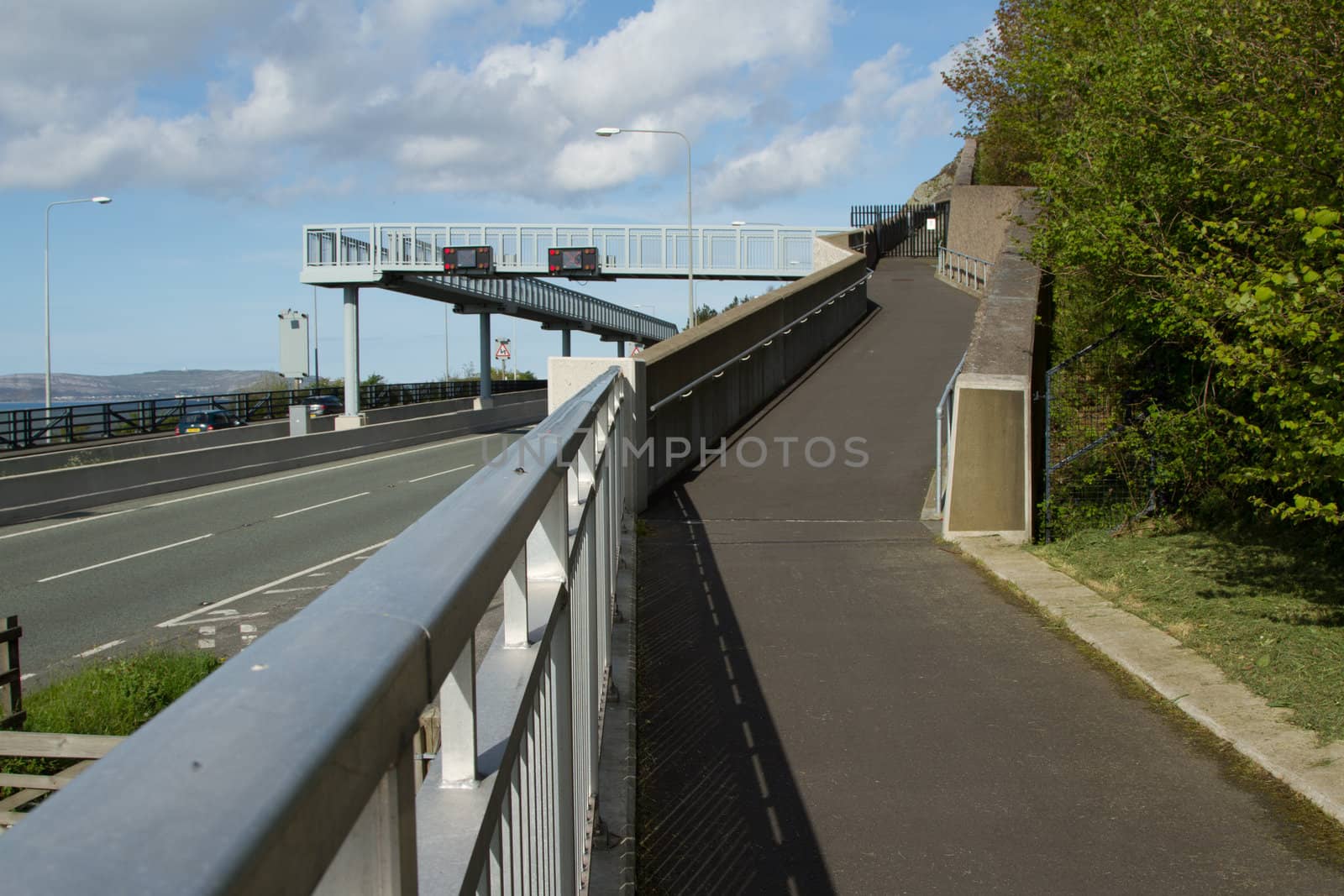 A footpath with metal railing leads to a footbridge with roadsigns over a road with a blue cloudy sky in the background.