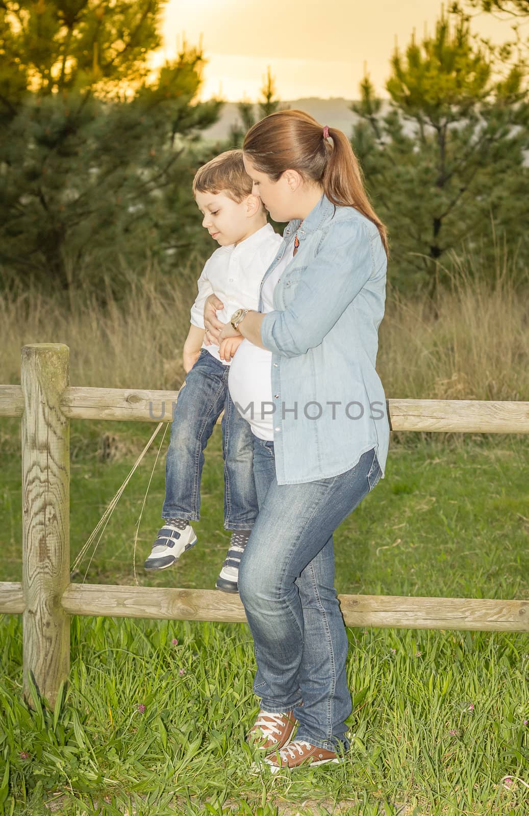 Pregnant mother sitting her son on a wooden fence in a field by doble.d