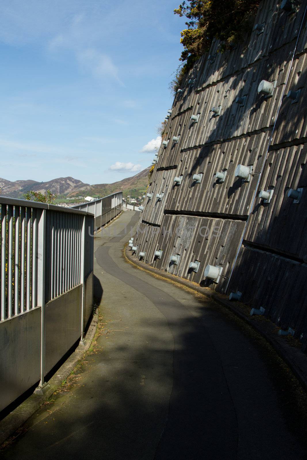 A footpath with railings winds along next to an engineered retaining wall with metal pins against a blue sky.