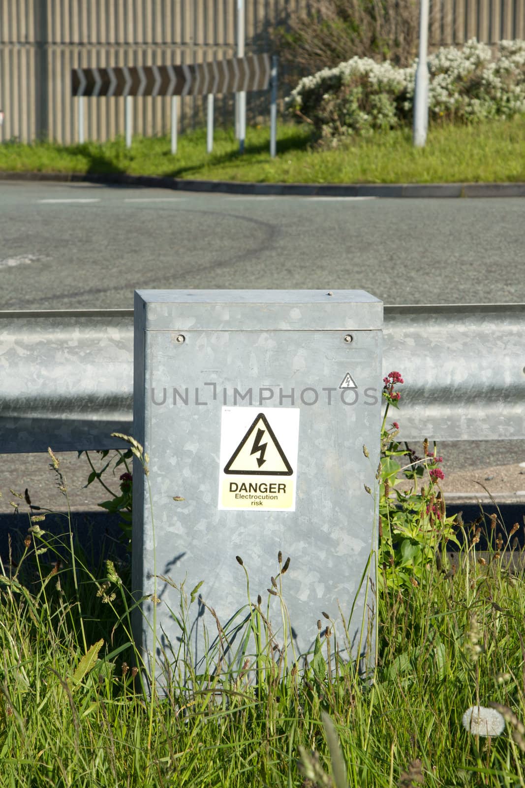 A sign for 'Danger' on a grey metal container in grass against a metal road barrier.