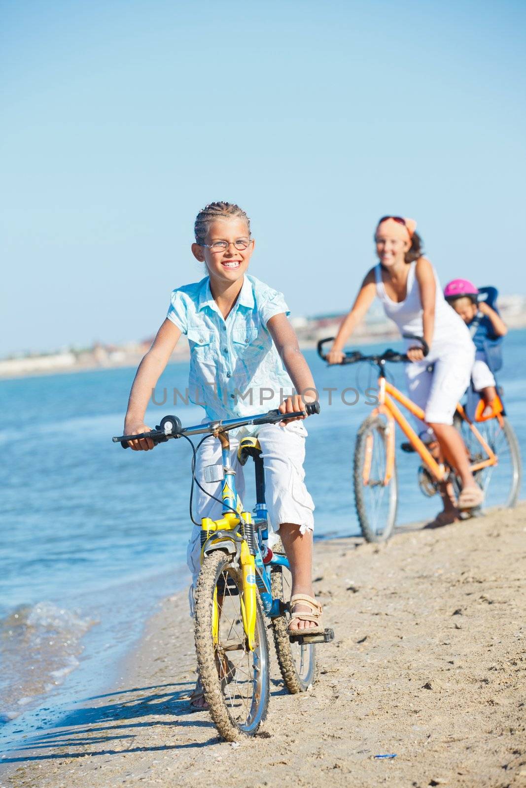 Cute girl with her mother and brother ride bikes along the beach. Focus on girl. Vertical view