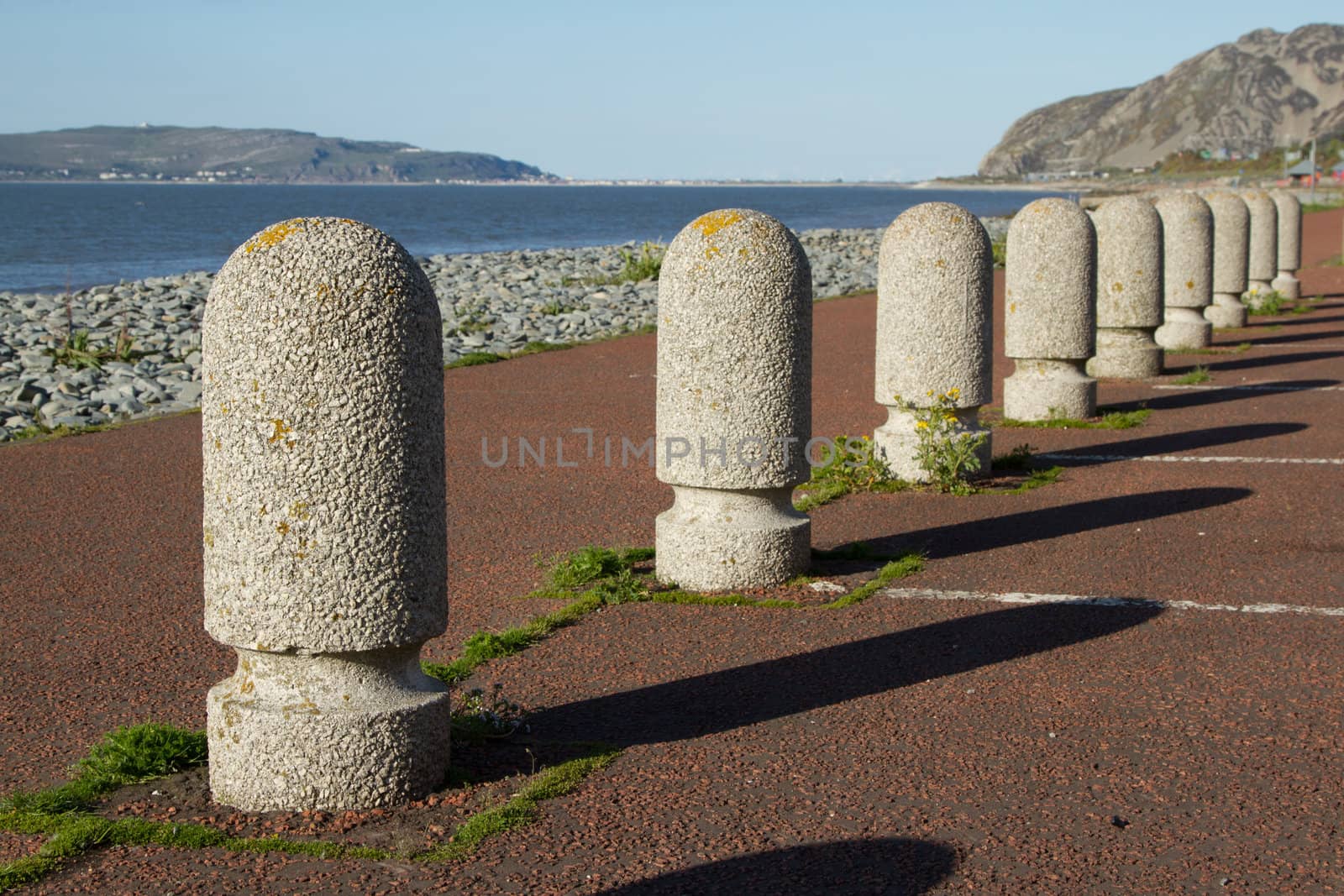 A set of concrete precast parking bollards stretch into the distance with a path, pebble beach and the sea in the distance.