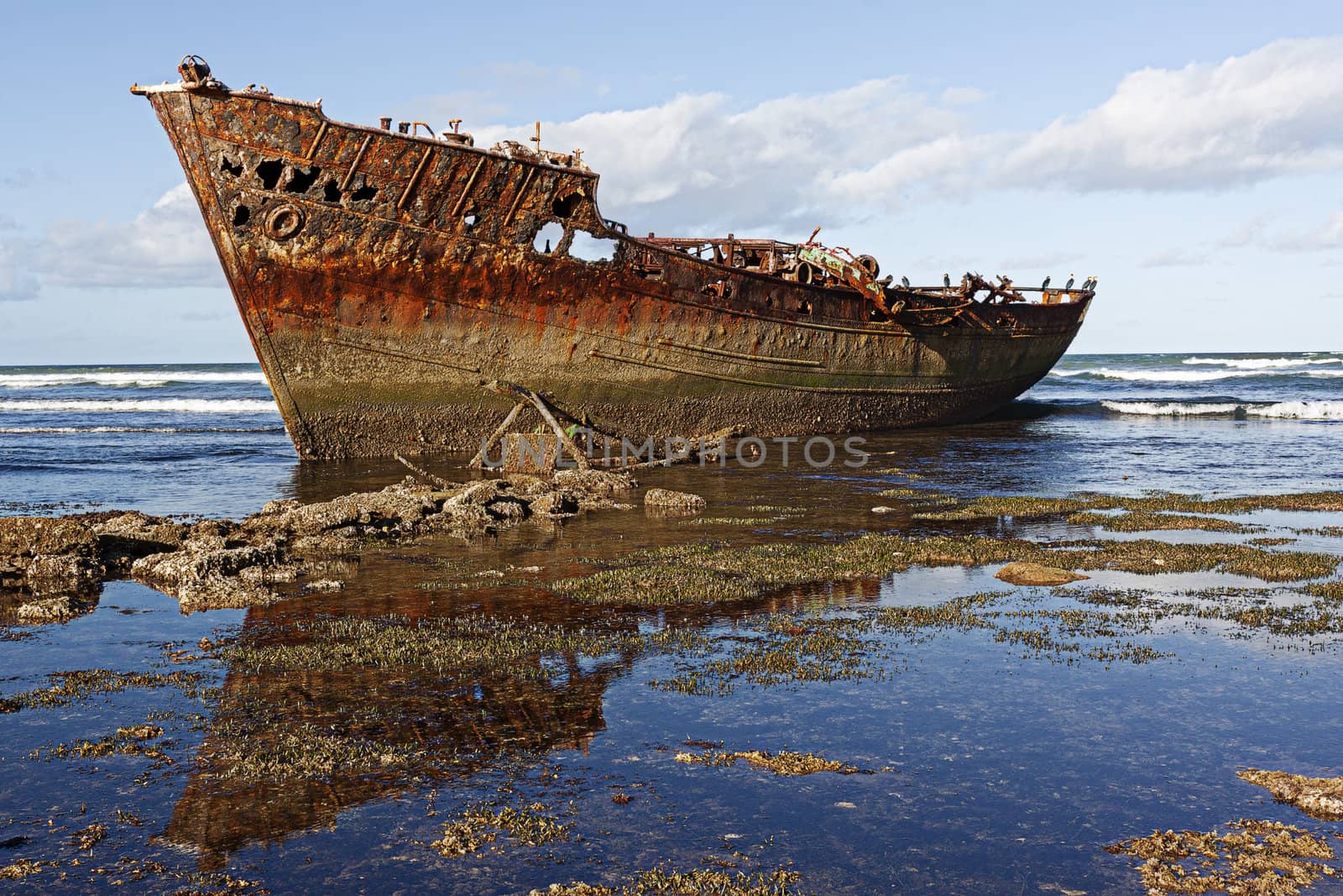 Rusty shipwreck on the rocks with blue sky and clouds in background.