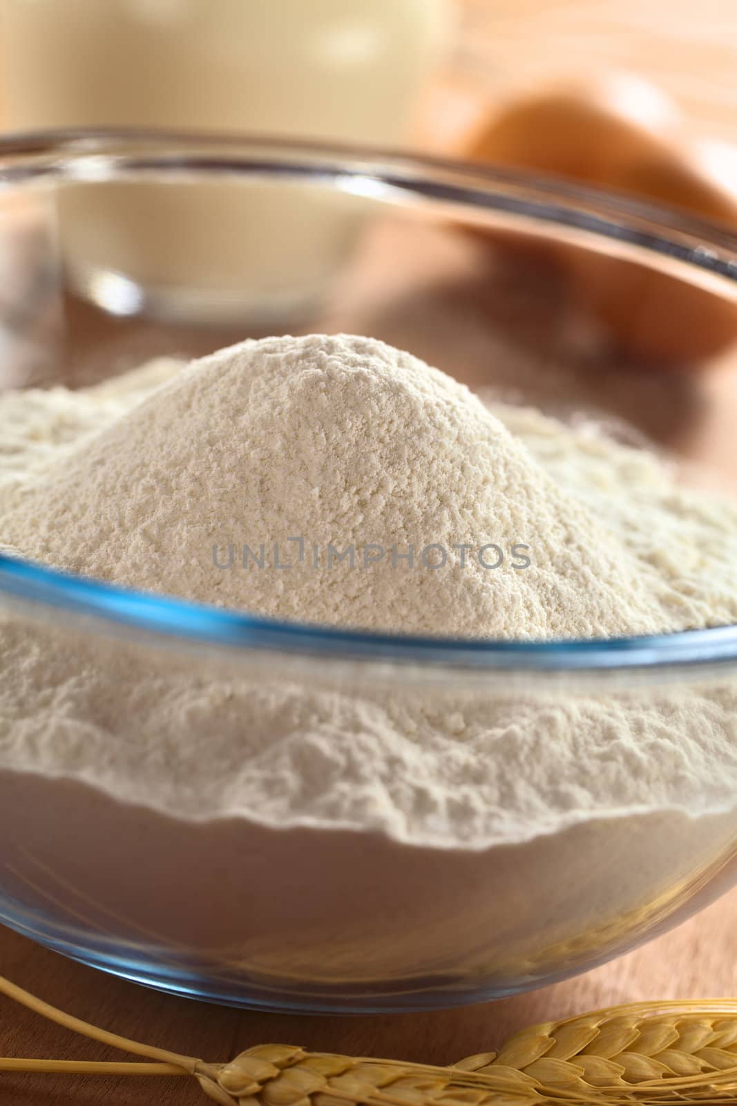 Preparing a dough for crepes or pancakes with wheat flour in glass bowl, milk and eggs in the back (Selective Focus, Focus on the top of the flour pile) 