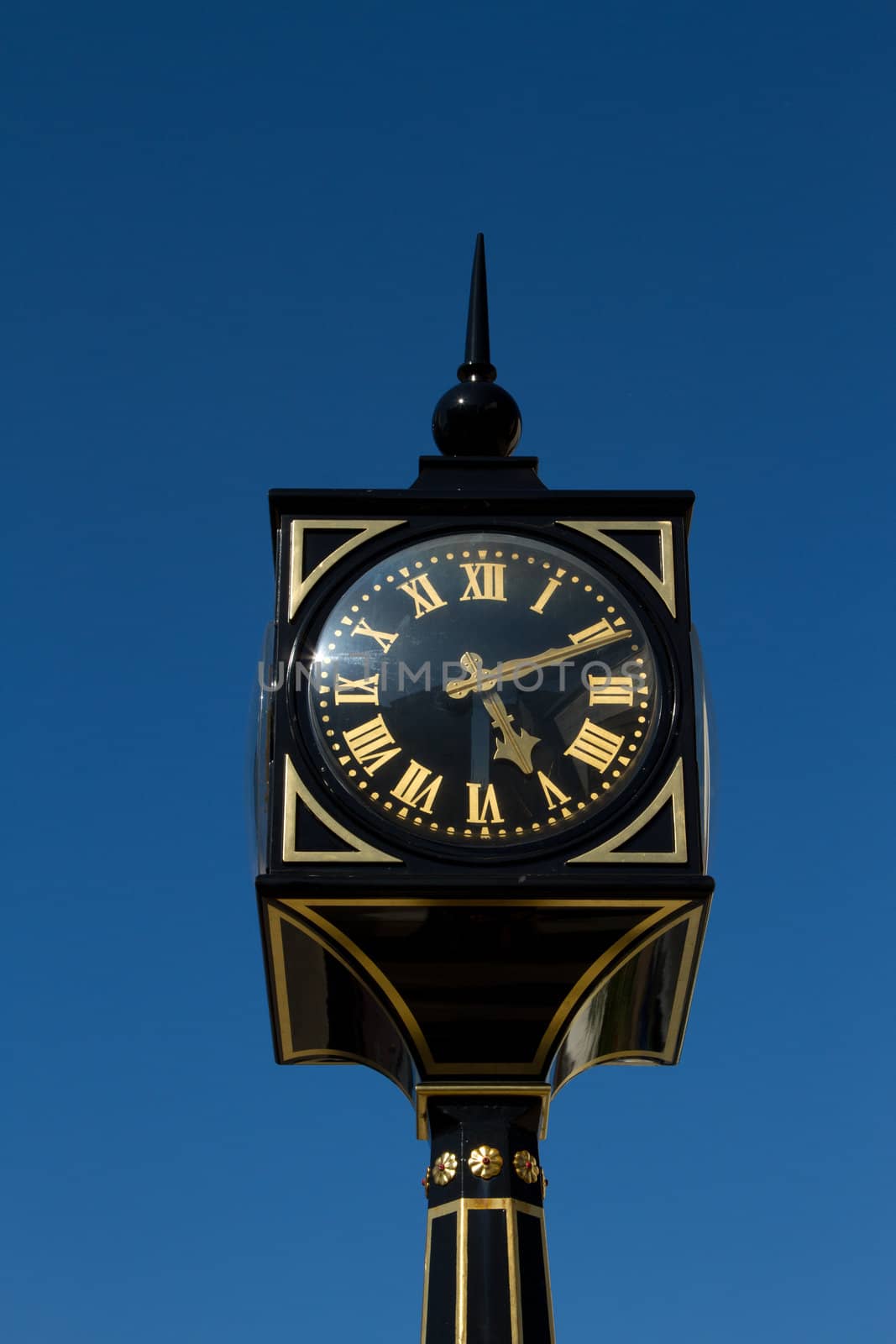 A clock with roman numerals on a post, pillar, painted black and gold with a clear blue sky in the background.