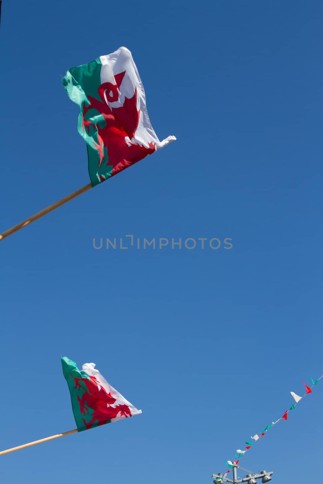 A pair of Welsh flags on poles fluttering and bunting in the distance against a clear blue sky.