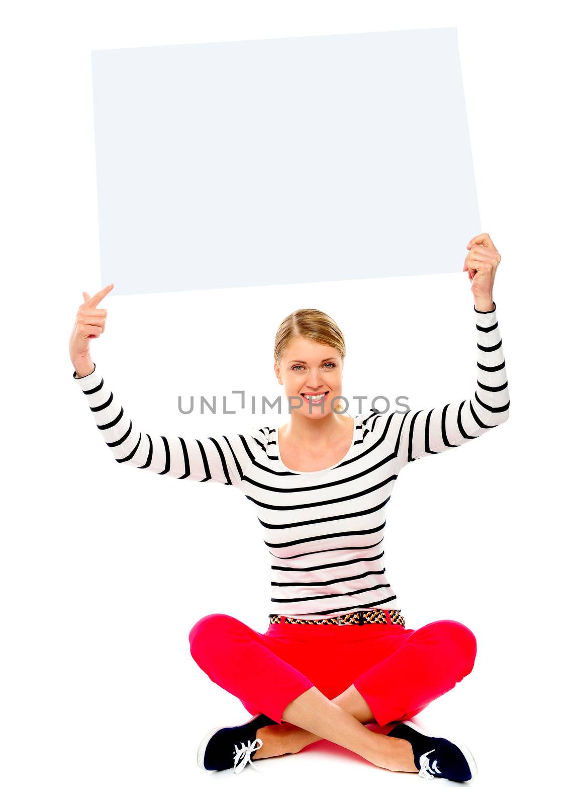 Beautiful young woman sitting on floor pointing towards blank banner ad over her head
