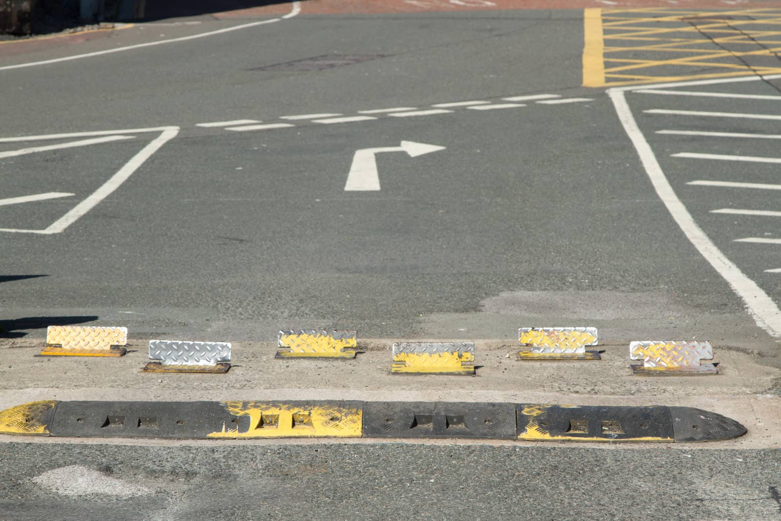 An exit with rumble strip, metal security blocks leading to a road with give way markings, a turn right arrow and various grid markings.