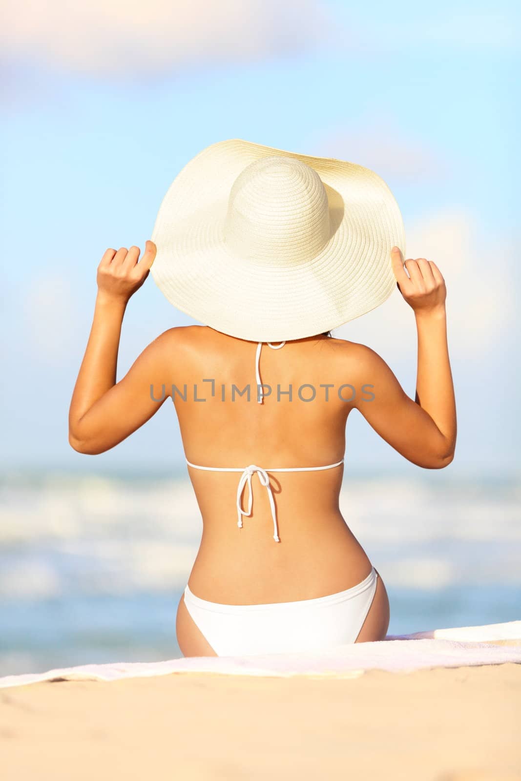 Travel vacation woman on summer holiday on beach holding her summer hat sitting in the sand enjoying ocean. Back view of beautiful girl in white bikini.