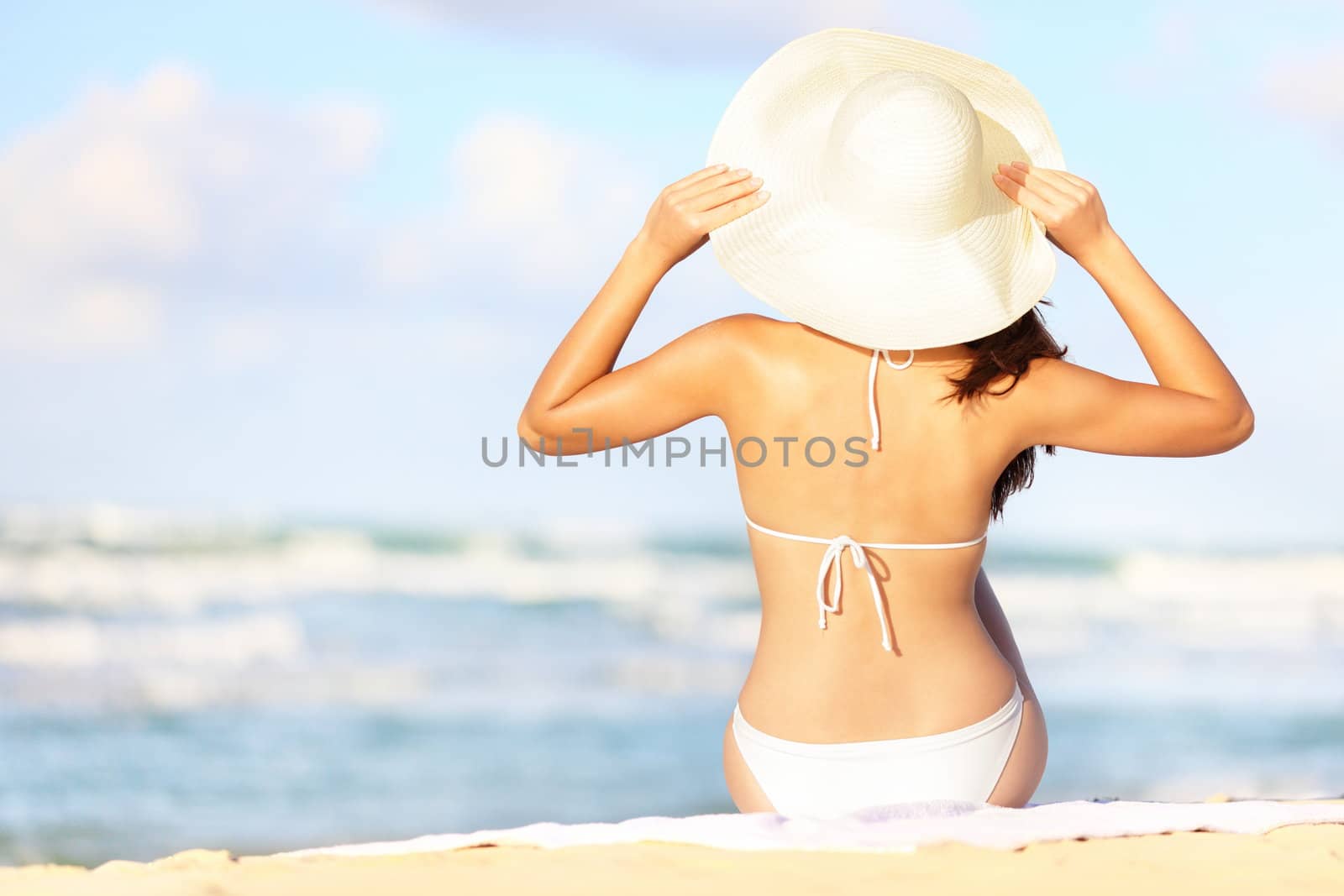 Summer vacation woman sitting on beach holding beach hat enjoying summer holidays looking at the ocean. Beautiful back side of model in bikini sitting down.