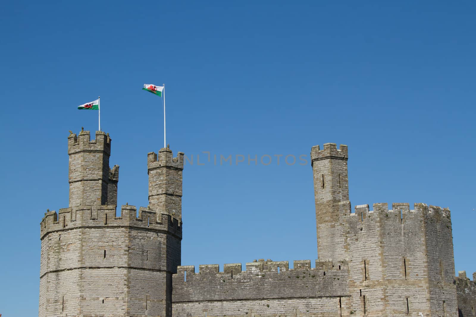 The towers, walls and ramparts of a castle with two Welsh flags flying against a clear blue sky.